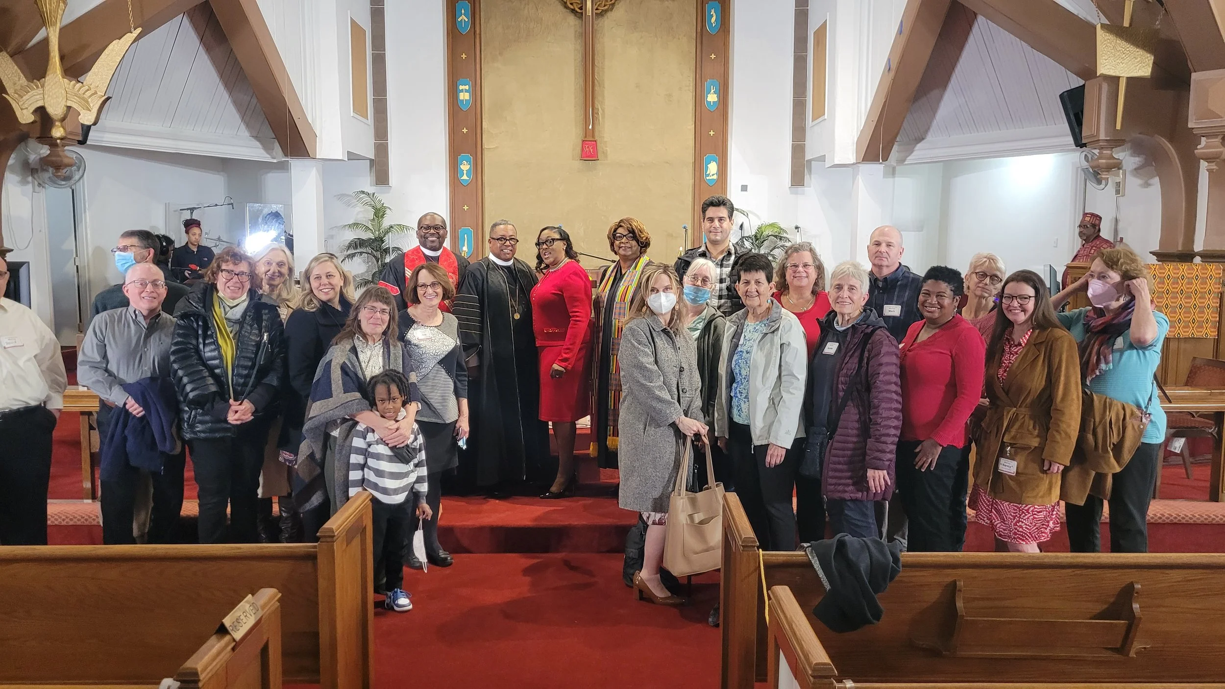 Group of diverse people gathered in a church, some wearing face masks, posing for a photo. The altar and wooden pews are visible in the background.