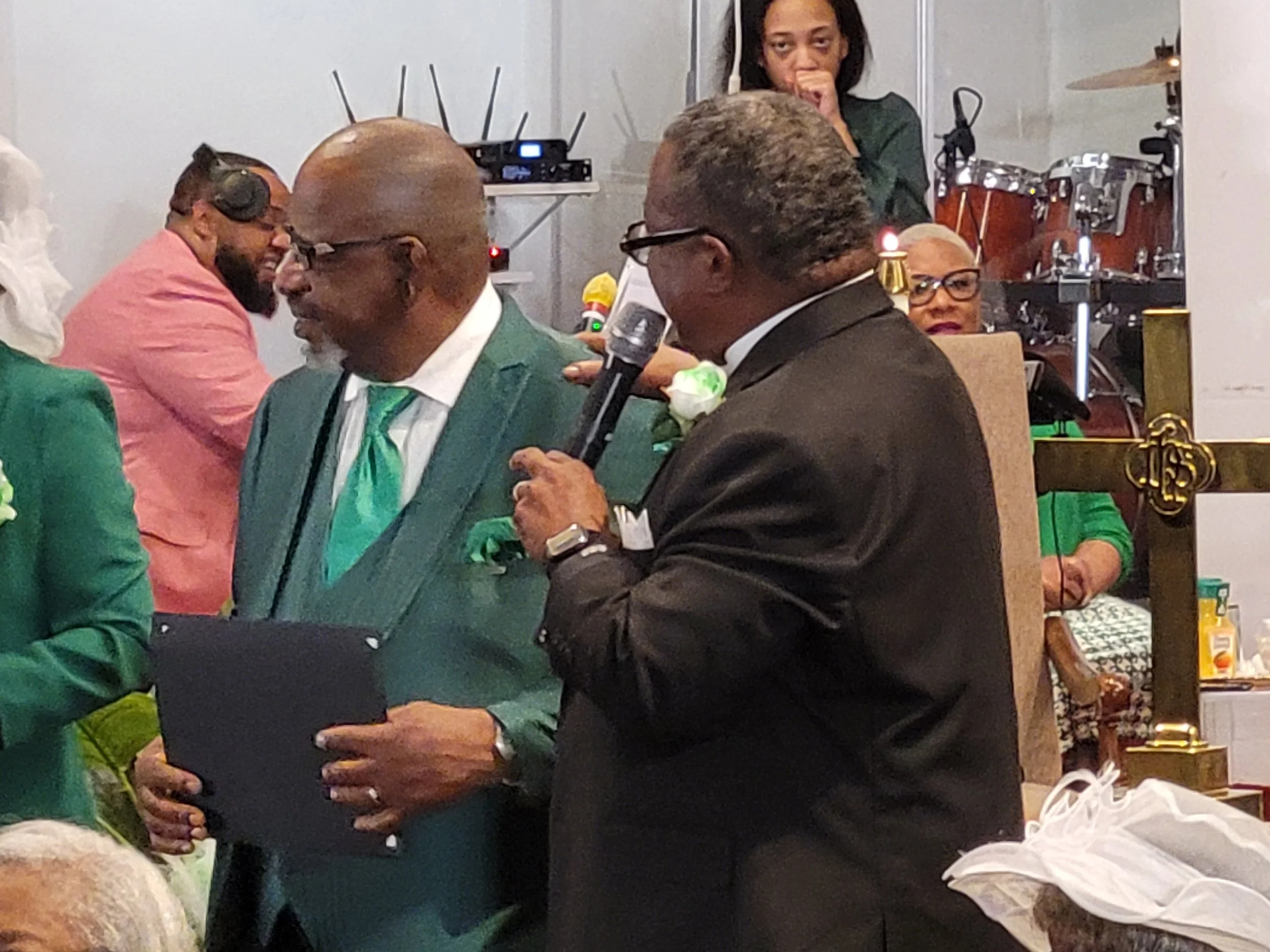 Man in black suit holding a microphone, speaking at a gathering, with older man in green suit and others in the background, some wearing glasses and green outfits.