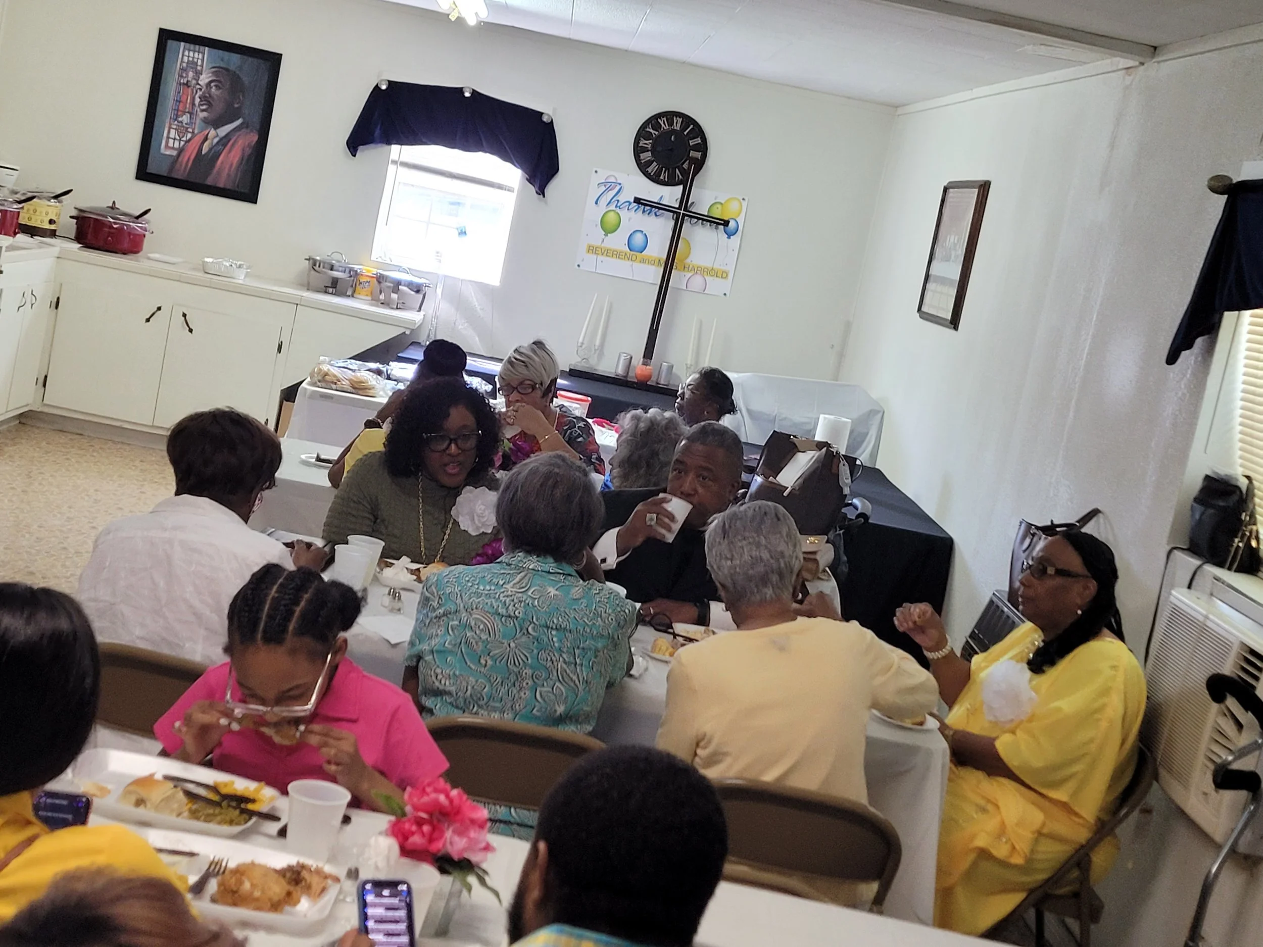 A group of African American people dining together at a celebration in a room with white walls and a window. They are seated around tables with food and drinks, some are talking or drinking, and one woman is feeding a child.