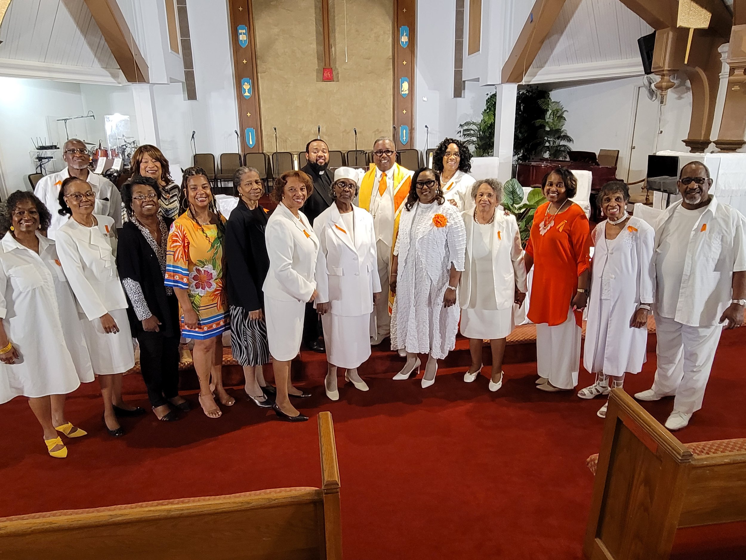 A group of diverse people, mostly women, standing together inside a church with a red carpet, smiling for a group photo. Some are dressed in white, with others in colorful outfits, and one man is in black clergy attire. The group is posed in front of