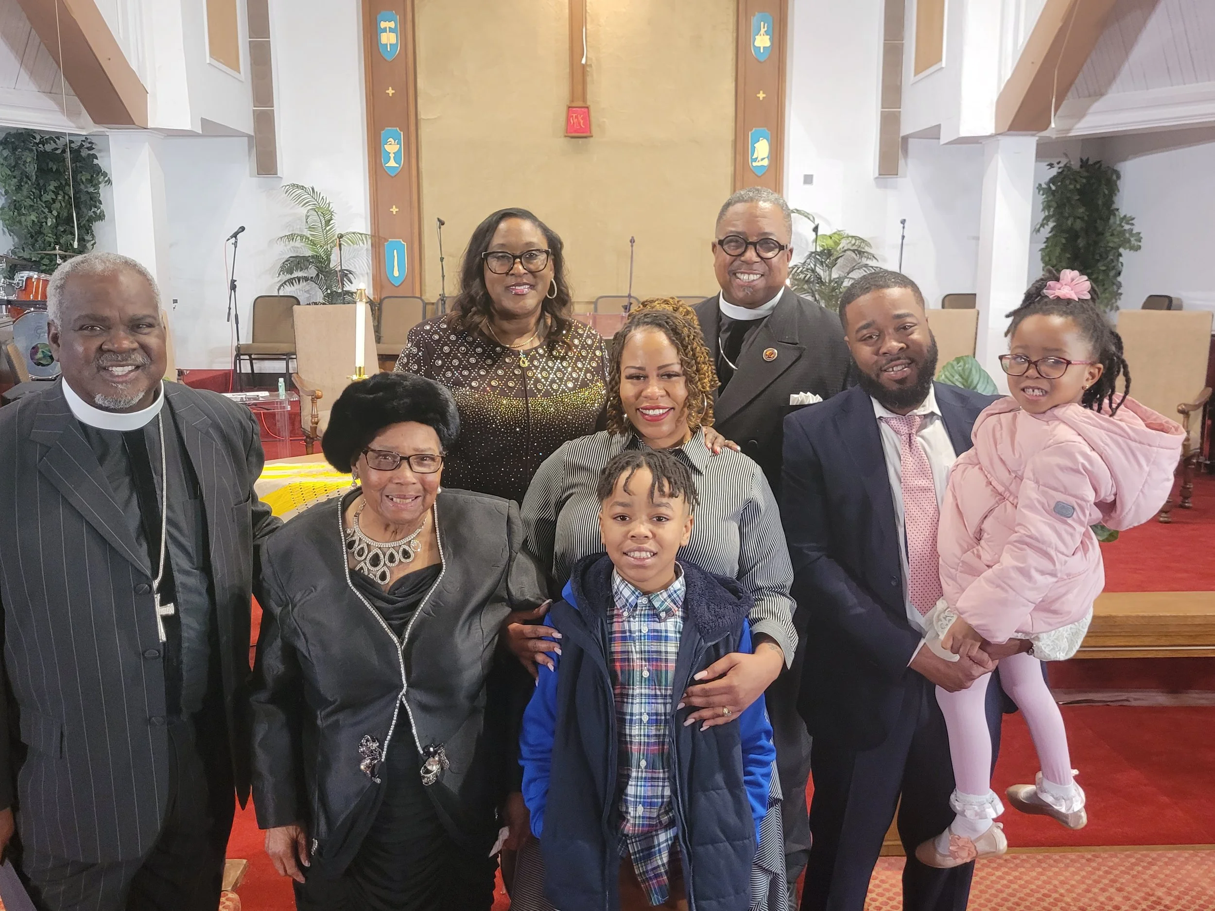 A group of ten diverse people, including children and adults, smiling in a church setting, with some wearing clerical attire and others in formal clothing.