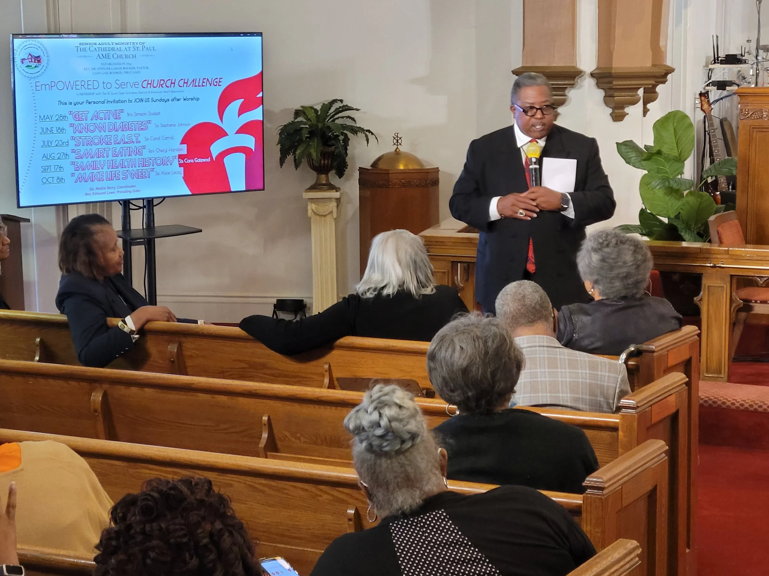 A man in a dark suit and glasses speaking into a microphone at a church event, with a group of seated women listening. A large screen behind him displays a presentation for a church challenge with dates and activities listed.