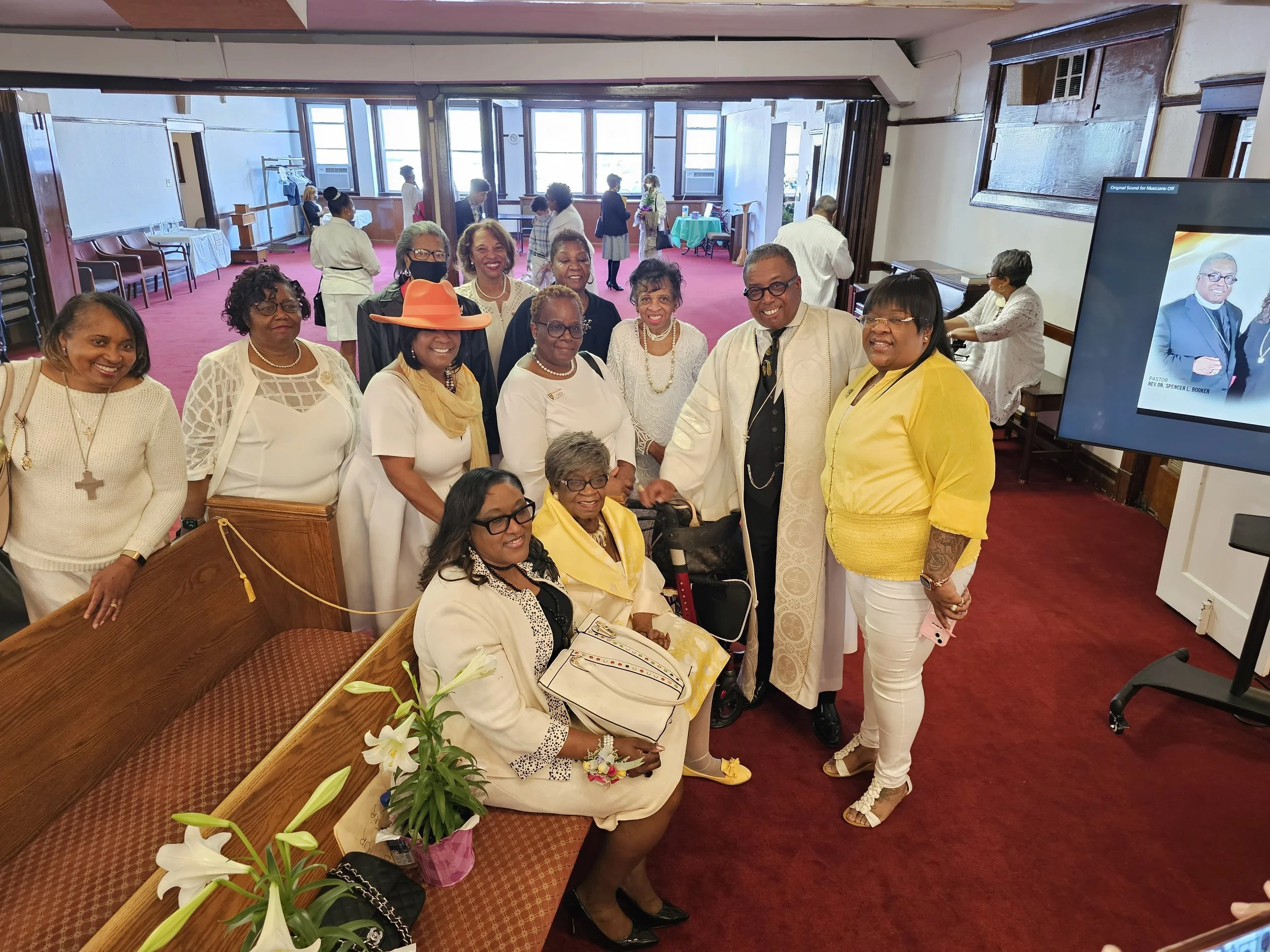 Group of people posing inside a church with red carpet, potted plants, and a large window in the background.