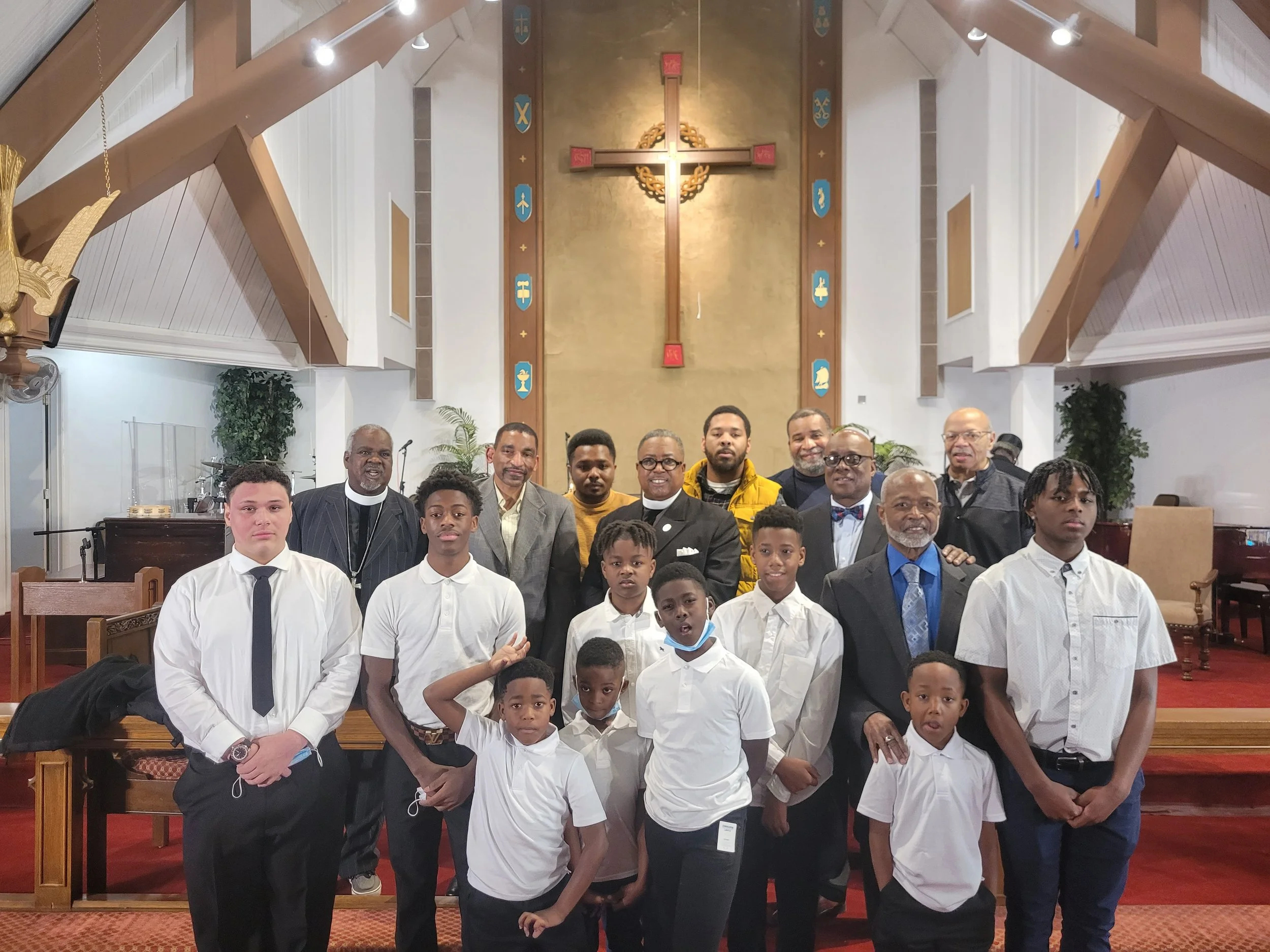 A diverse group of boys and men standing inside a church with a gold cross and religious symbols behind them, some dressed in formal and others in casual attire.