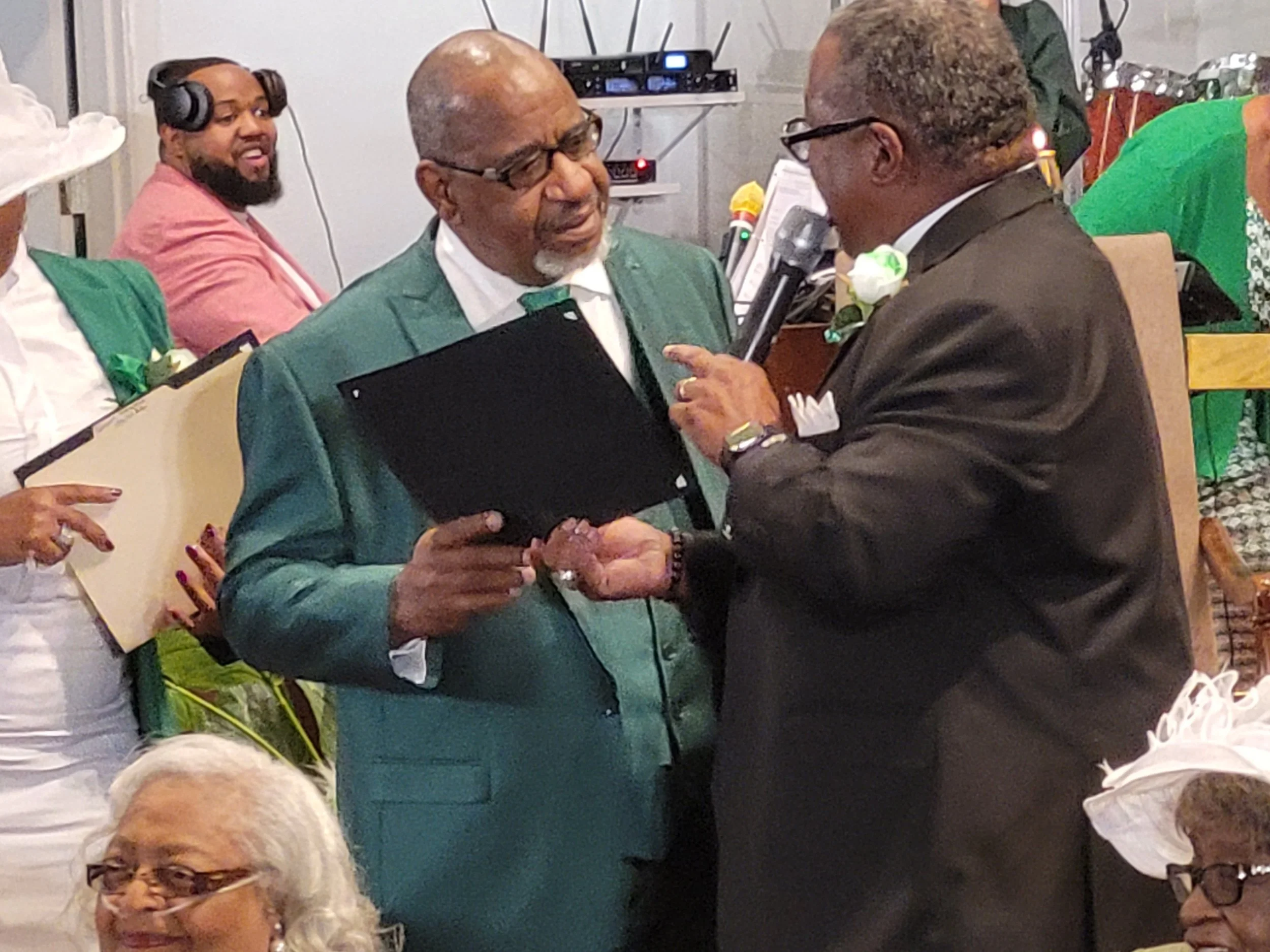 Two men in suits, one older with glasses and a gray beard, the other younger, engaged in a conversation at a formal event, surrounded by seated attendees.