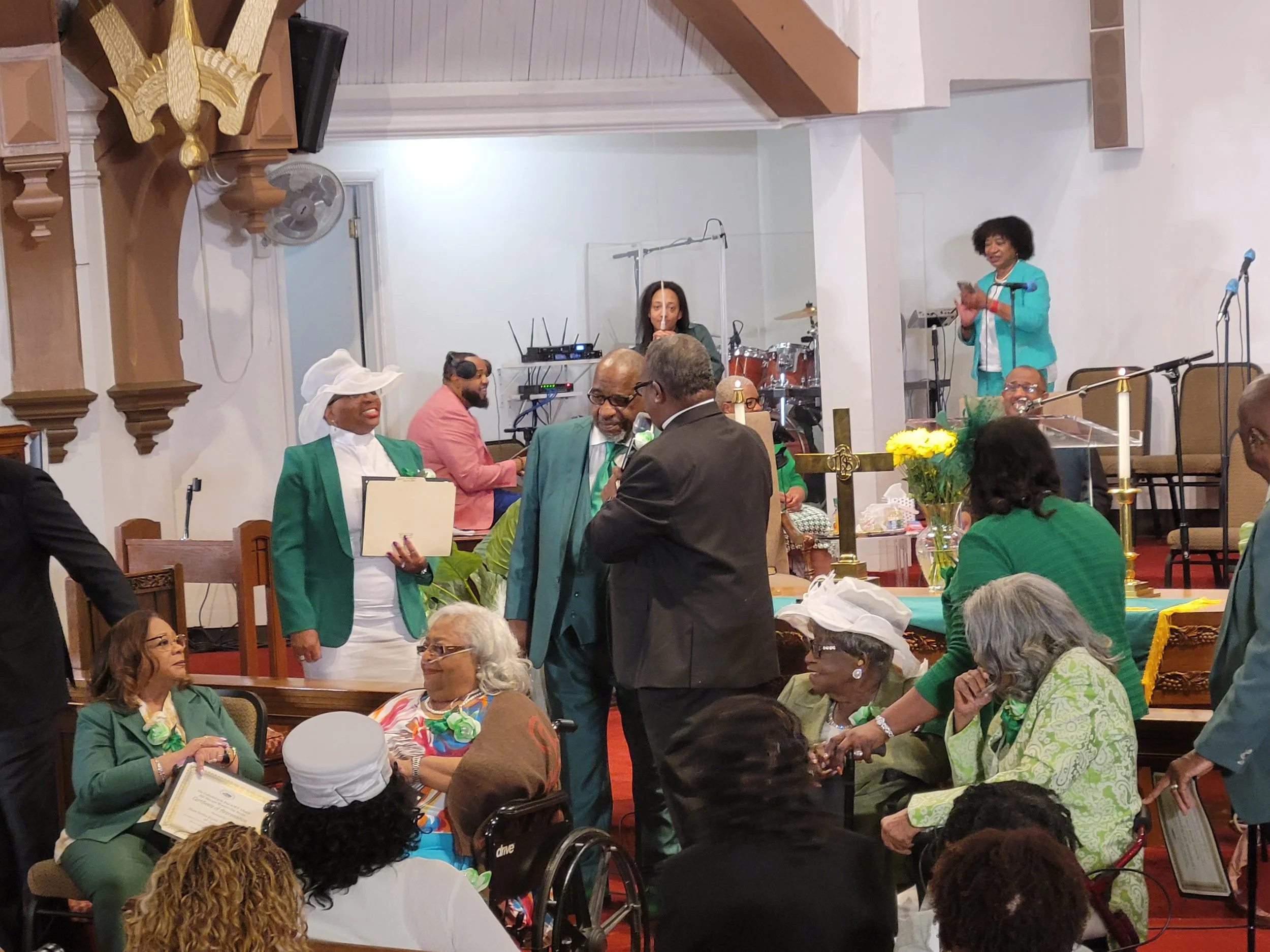 A religious or community gathering inside a church with people of all ages dressed in formal green attire. Some individuals are seated, while others stand, engaging in conversation. There is a person speaking into a microphone and a band playing in t