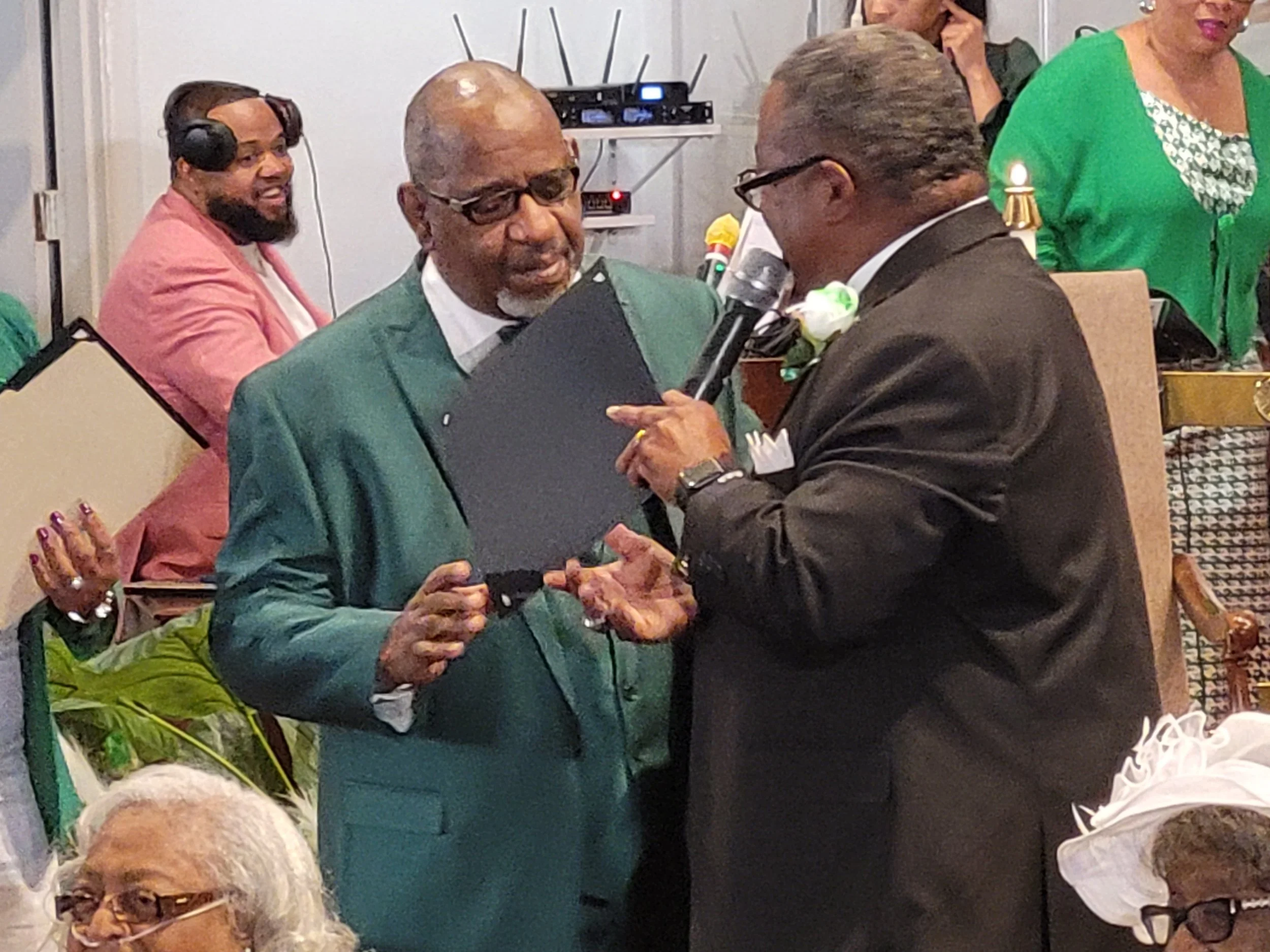 Two men are engaged in a conversation at a formal event, one holding a microphone and a folder, and the other wearing glasses and a boutonniere. Several other people are visible in the background and foreground.