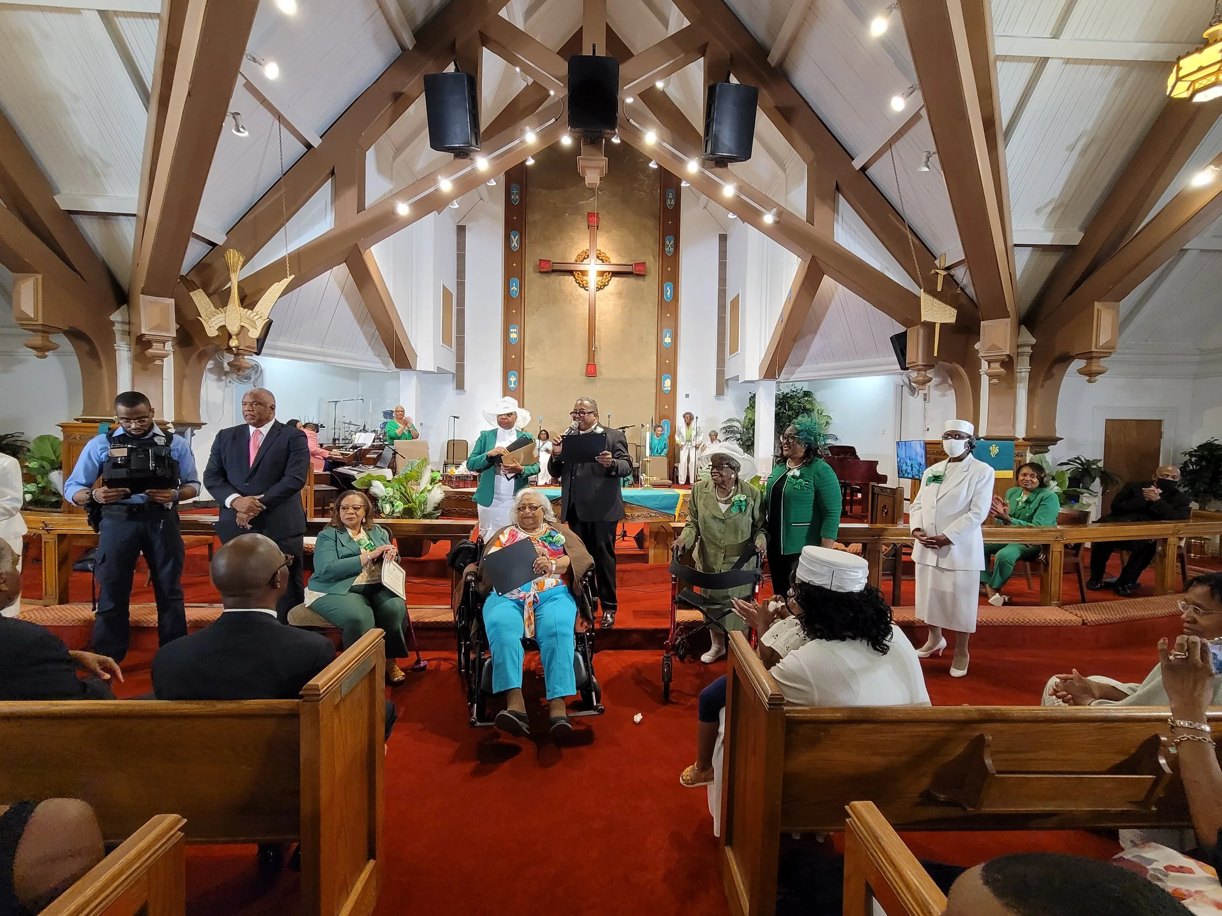A group of people gathered in a church during a ceremony; some are standing on the altar and others seated in the pews, with a large cross hanging on the wall behind them.