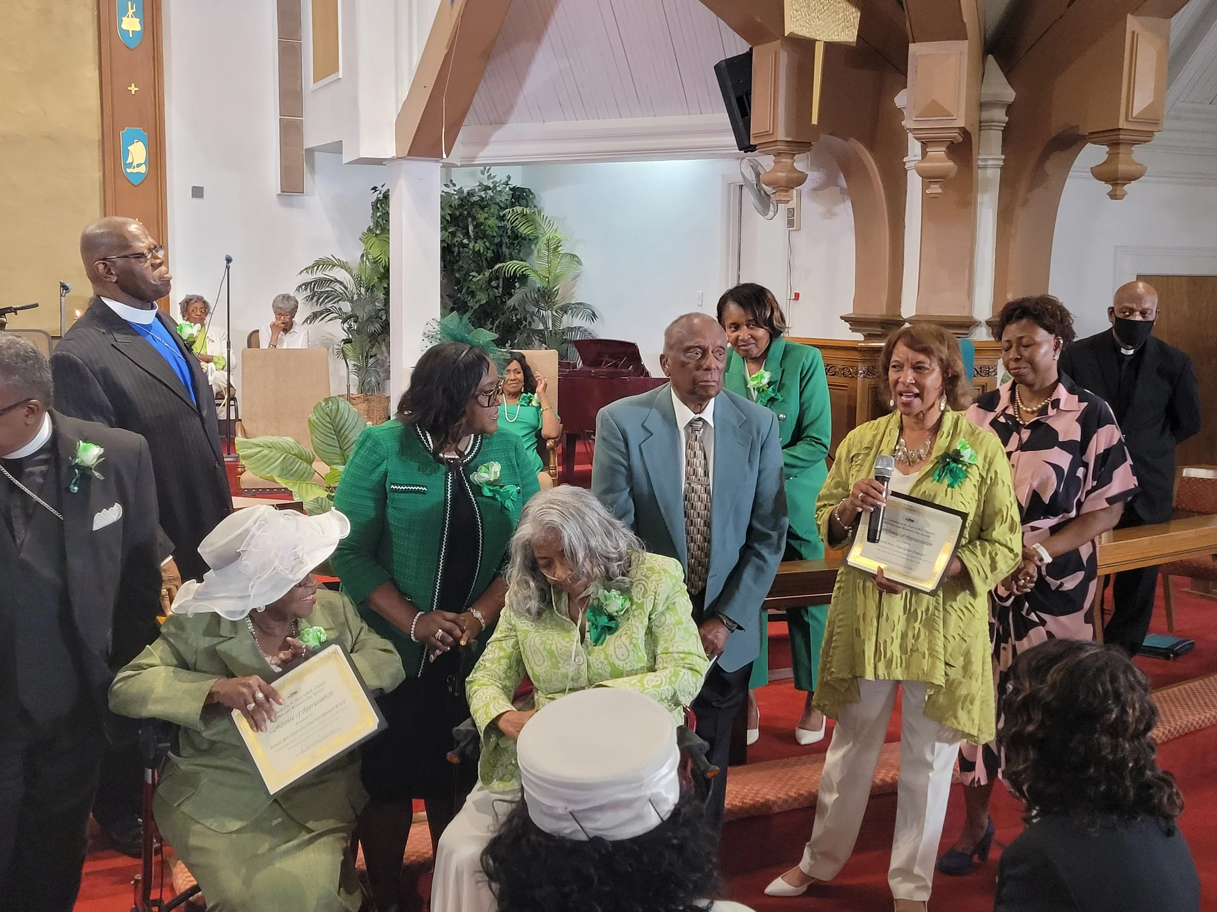 Group of people gathered in a church, some holding certificates, celebrating an event or award ceremony. The woman in yellow is speaking into a microphone. There are decorated with green ribbons and plants in the background.