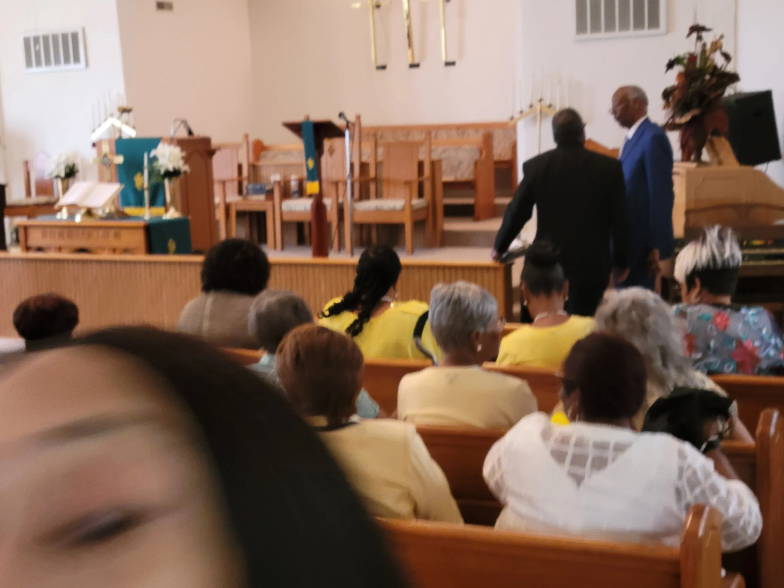 Inside a church with people seated in pews, some facing the front where a stage with chairs, flowers, and religious symbols is visible. Three men are standing near the stage, engaged in conversation.