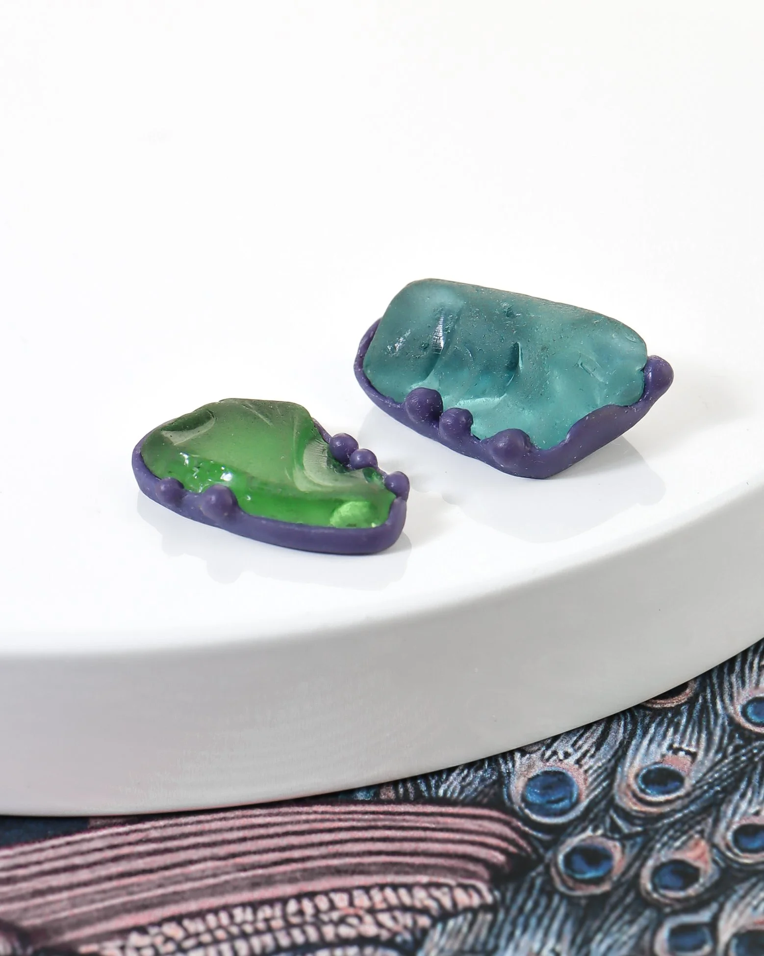 Close up image of two pieces of sea glass, green and aquamarine, in settings carved from purple wax with droplet detail to secure the glass