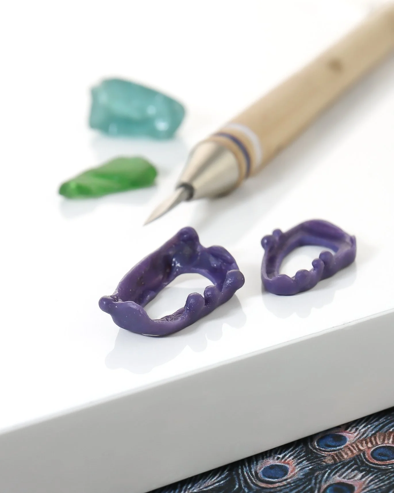 Settings carved from purple wax, sitting on a white acrylic sheet with a carving tool, and two pieces of green and aqua coloured sea glass in the background
