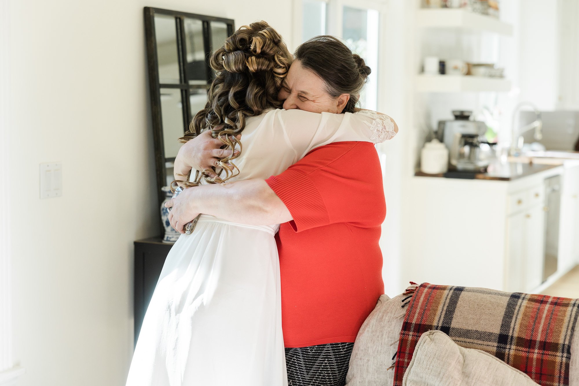 woman with long curly hair wearing white robe hugs woman wearing red tshirt