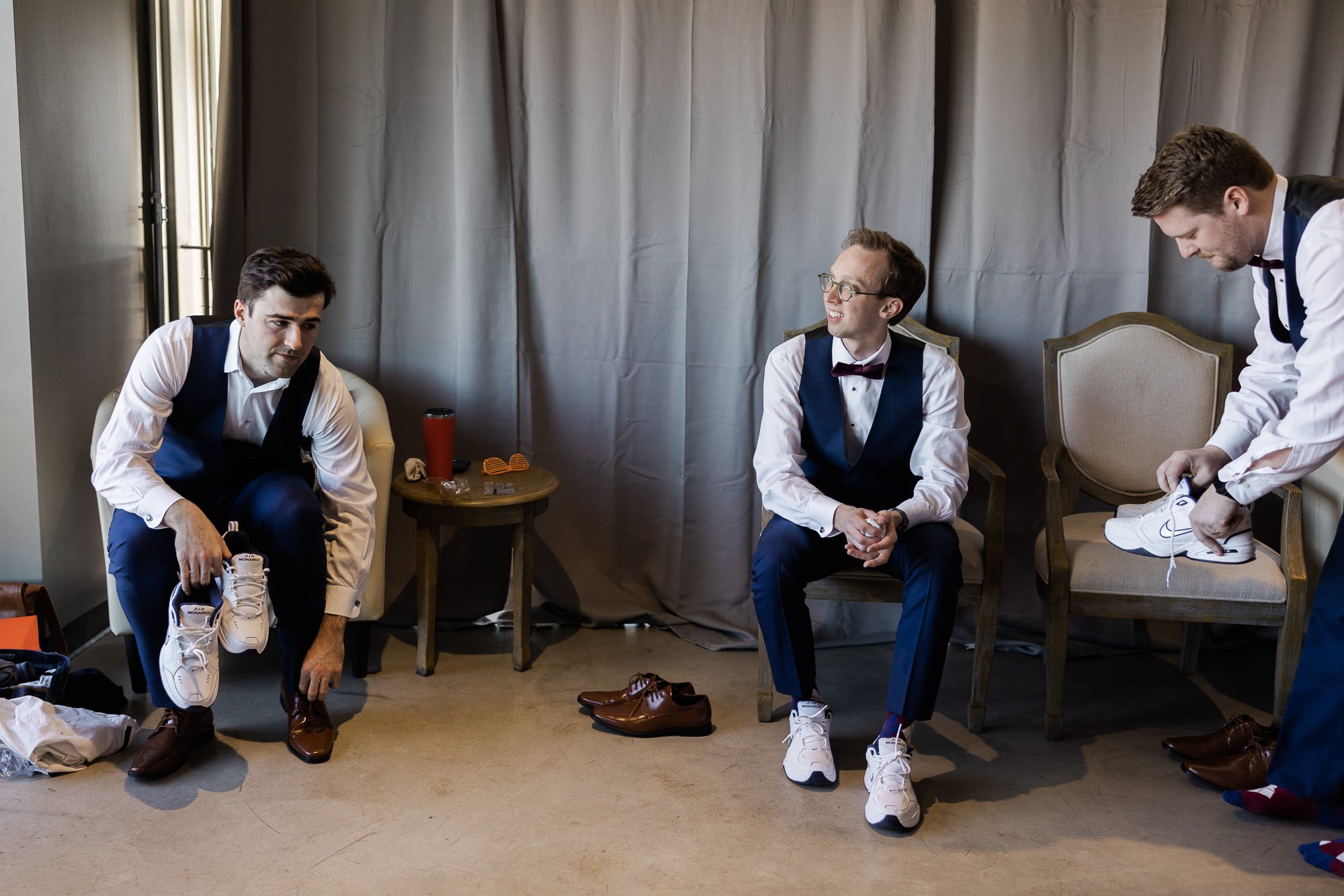 two men in white shirts and blue pants sit while putting on tennis shoes as a third man stands and ties shoe on chair