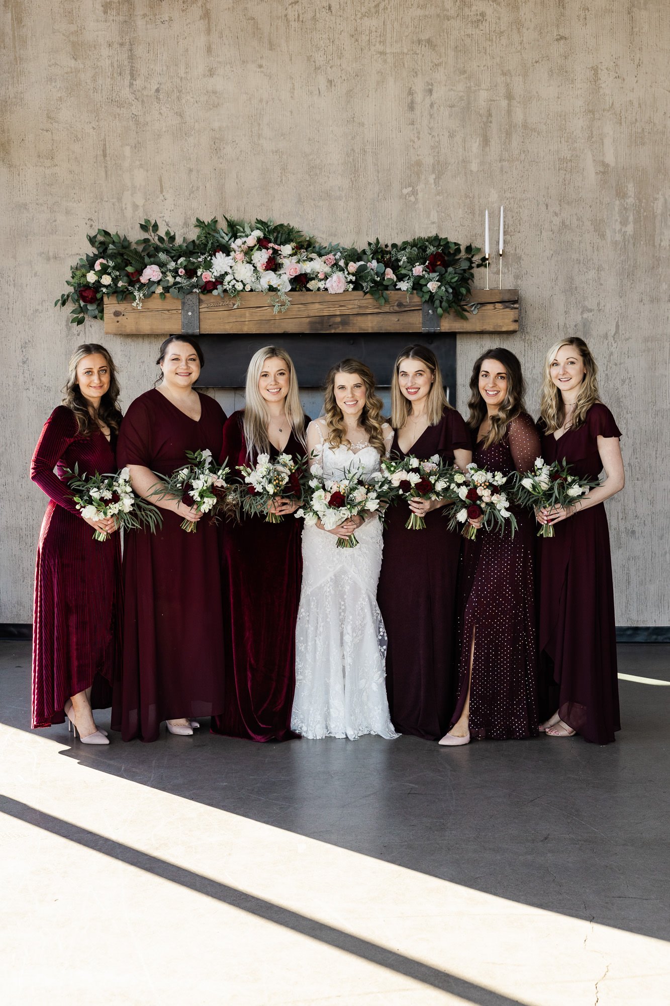 six women in burgundy dresses and a woman in a white wedding dress stand in front of fireplace mantel at the black pearl on the columbia