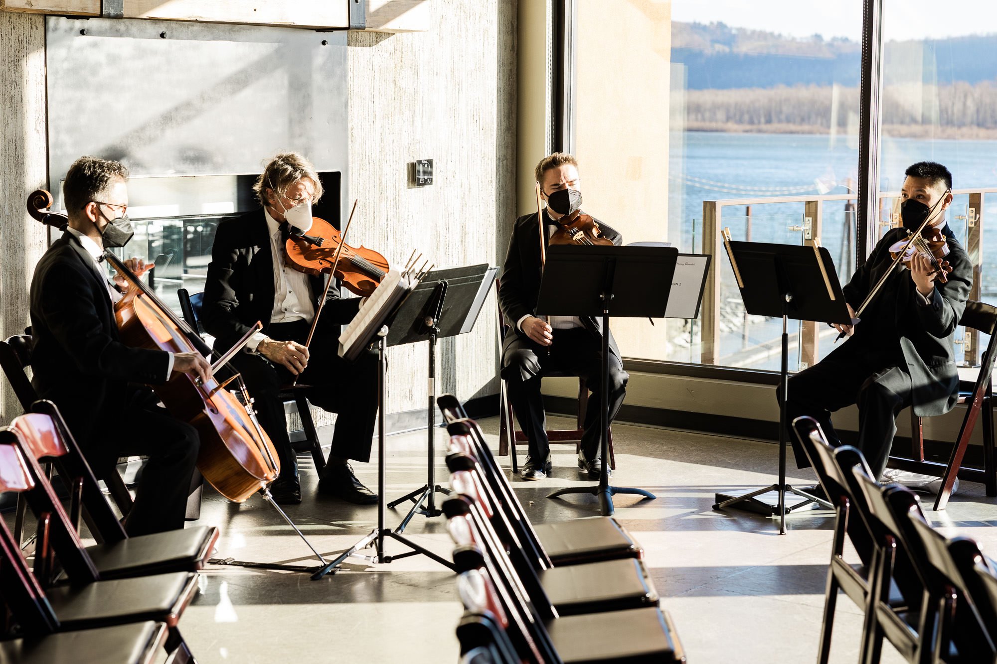 string quartet plays instruments in front of large windows at wedding ceremony