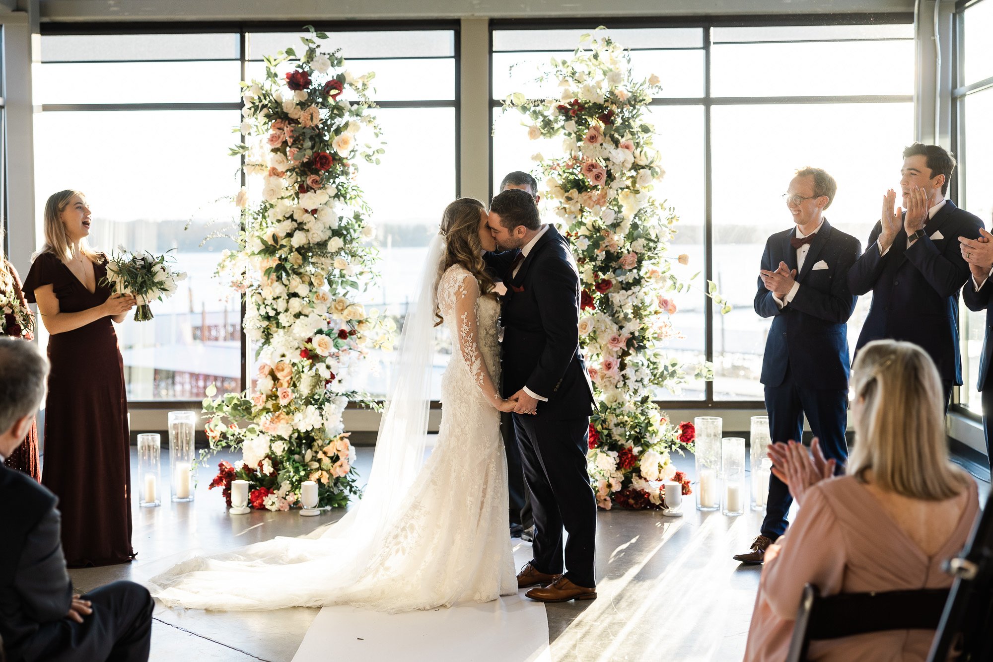 bride and groom kiss in front of floral arbor and large windows during wedding ceremony at black pearl on the columbia
