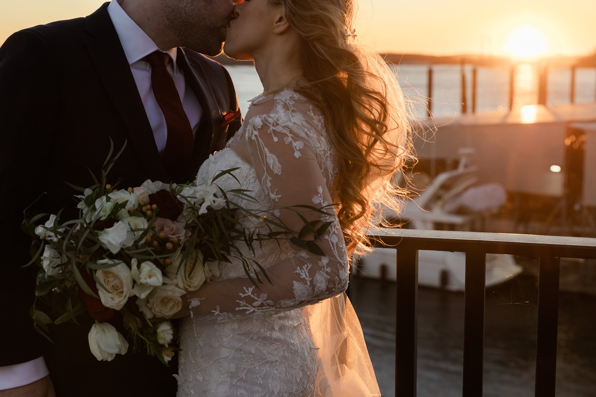 bride holds bouquet of flowers while kissing groom in front of sunset