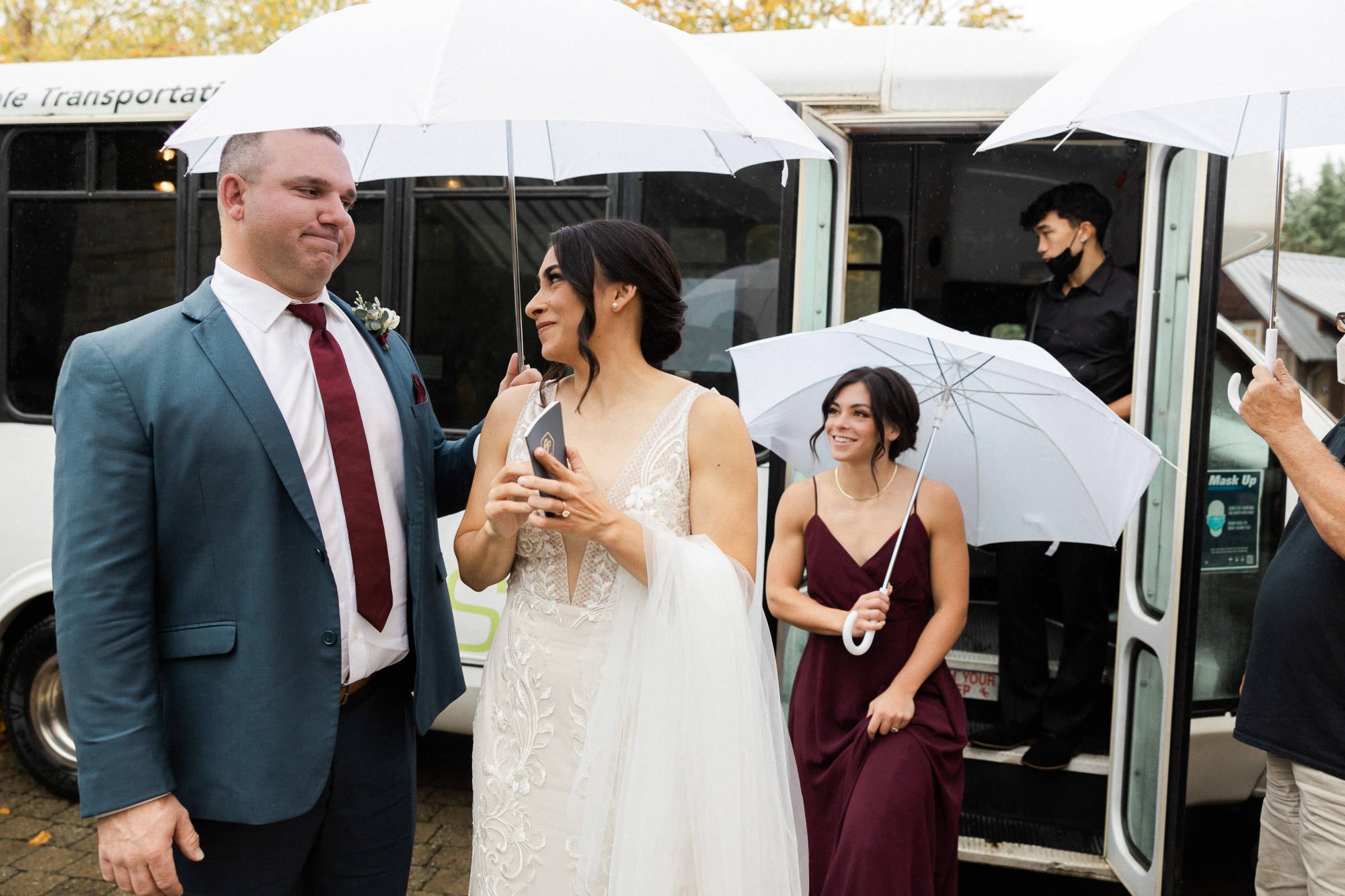 woman in burgundy dress holding white umbrella exits bus behind bride in white dress and groomsmen in navy blue suit holding white umbrella