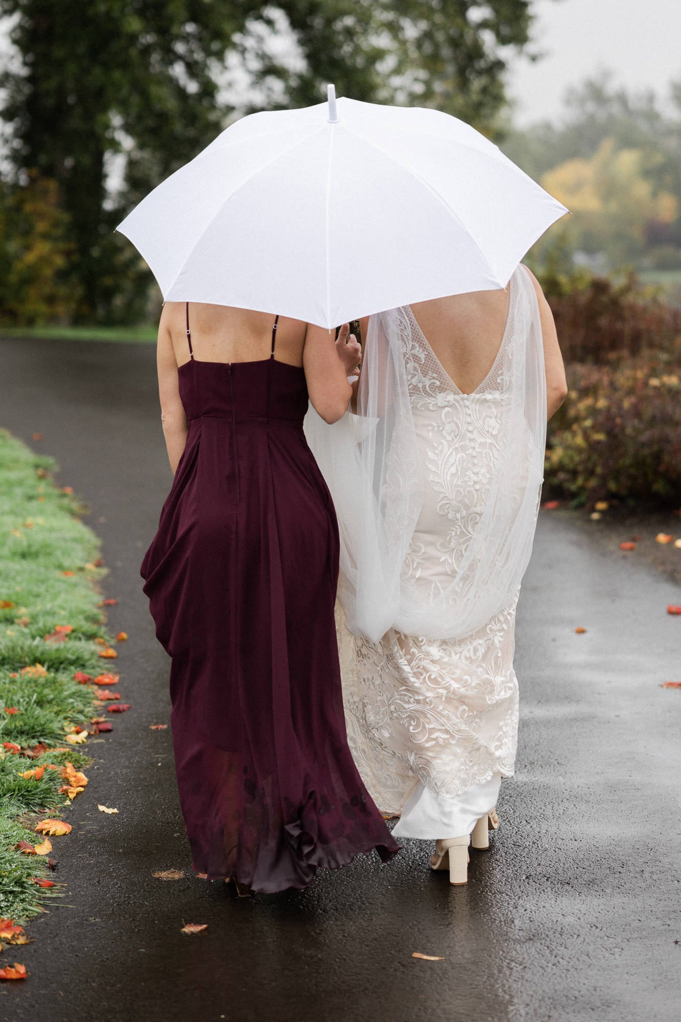 a woman in a burgundy dress and a bride in a bridal gown walk away while holding white umbrella in lake oswego, oregon
