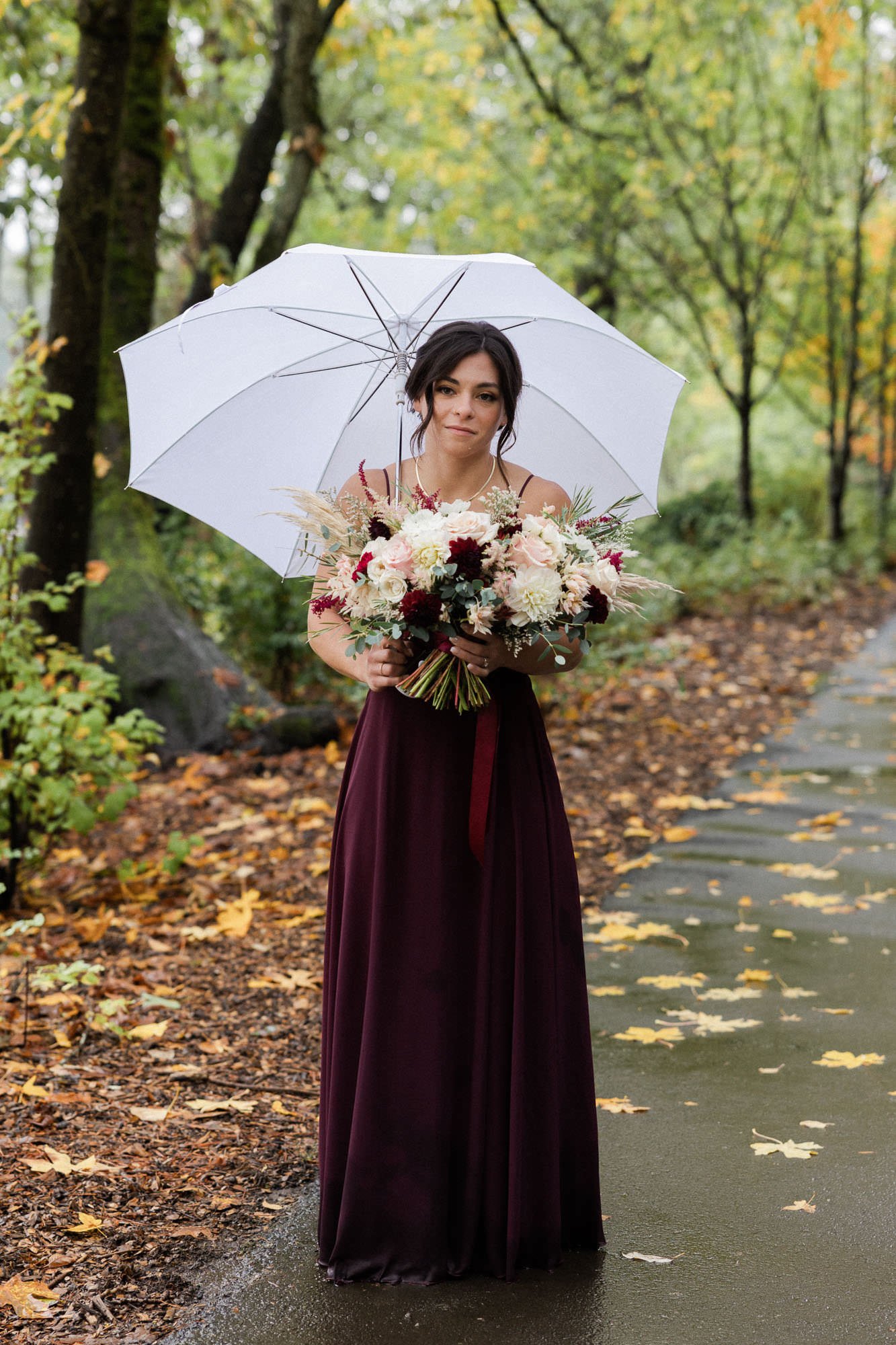 woman in burgundy dress holds floral bouquet and white umbrella at lake oswego park