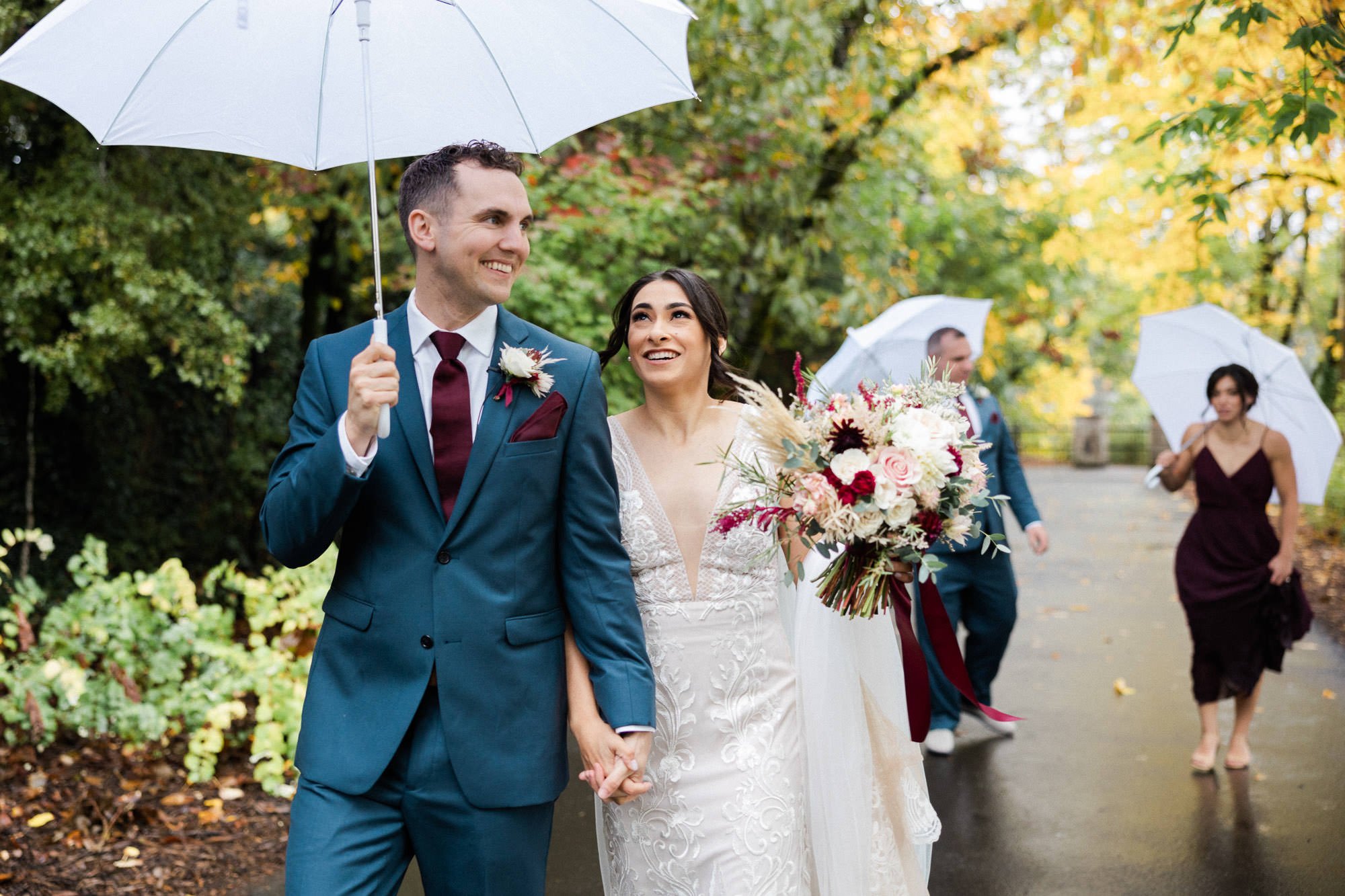 bride and groom walk through park holding white umbrella in front of bridal party