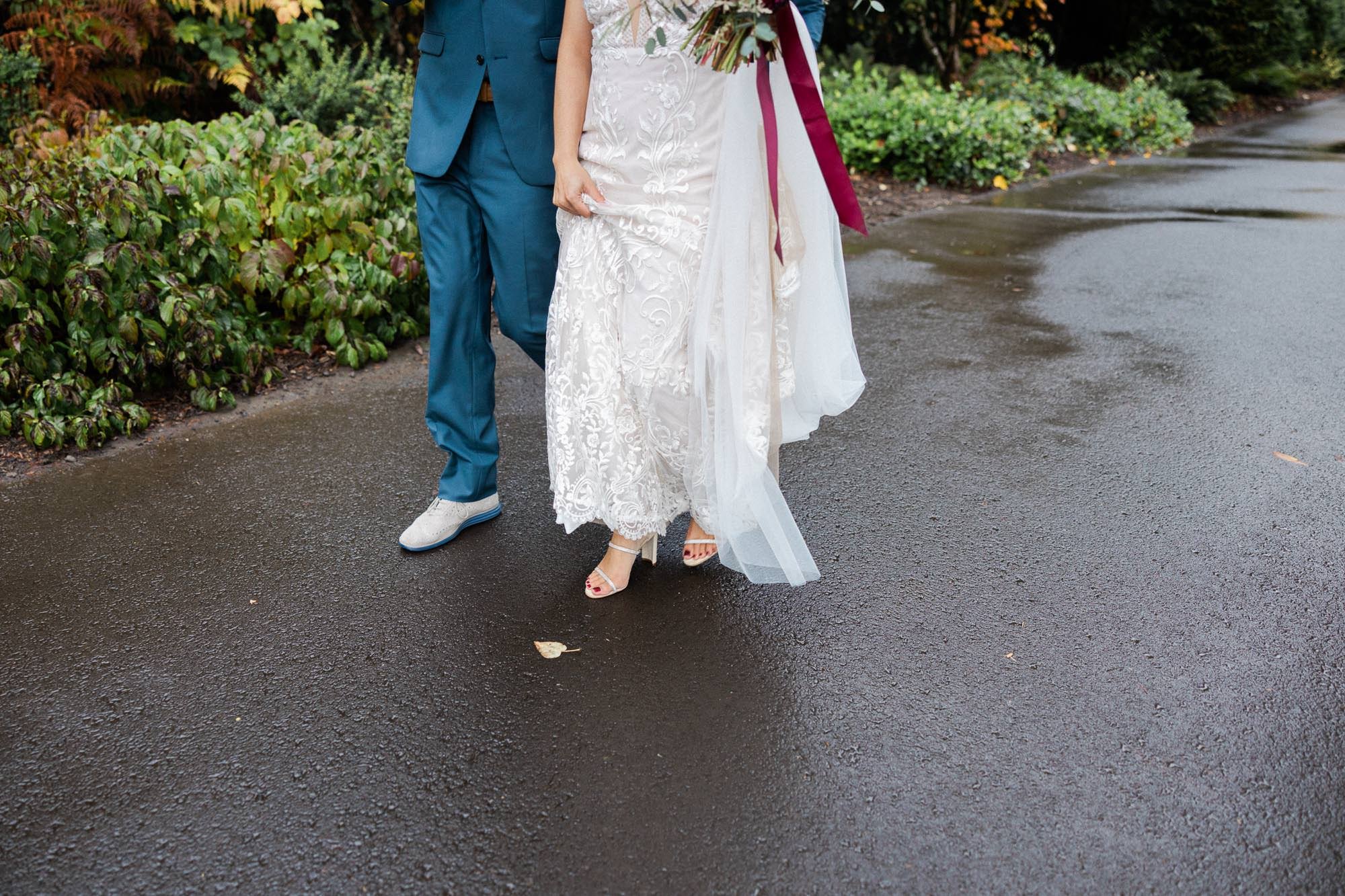legs and feet of groom in navy blue suit and bride in white wedding dress walk on wet asphalt