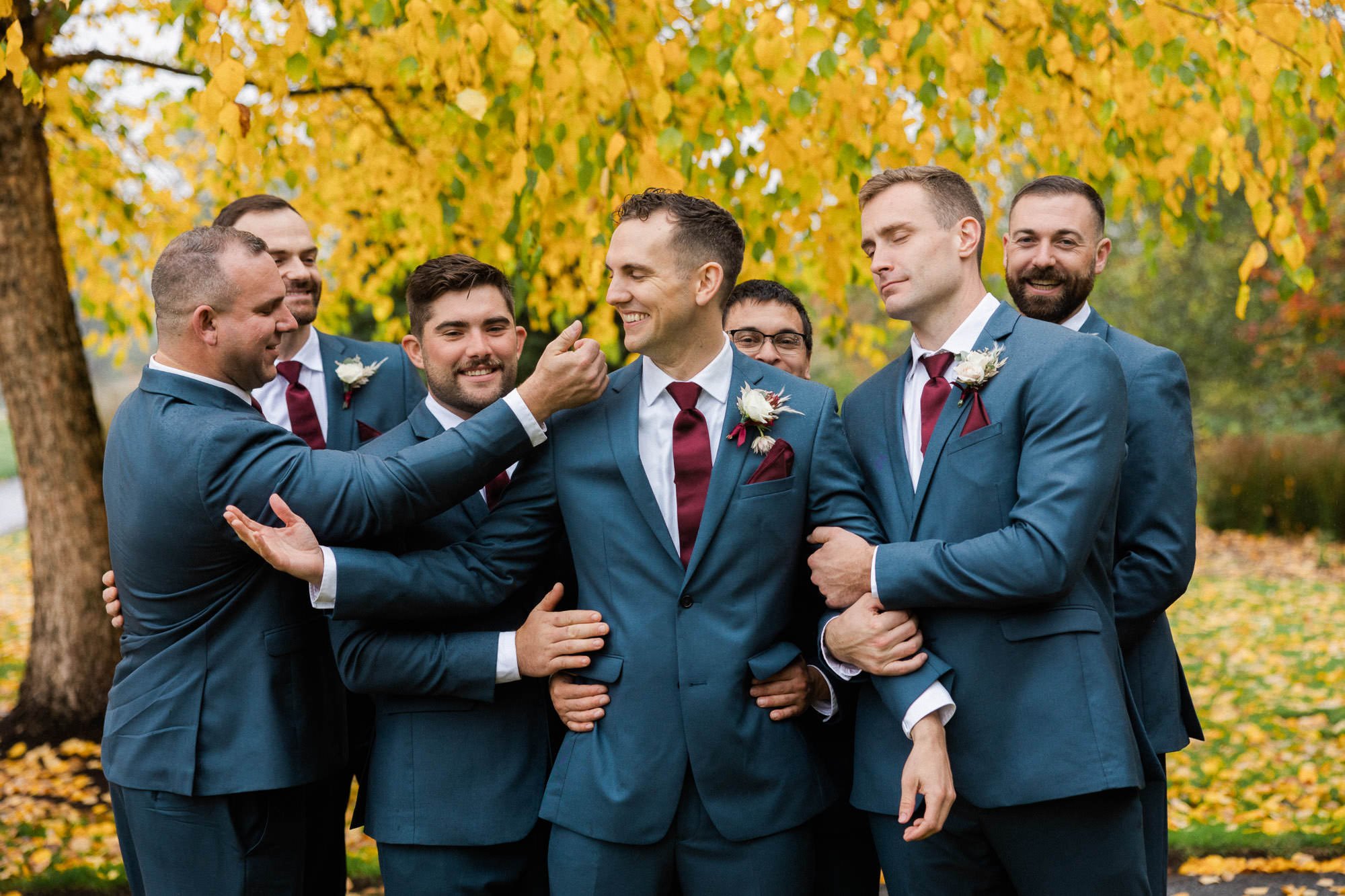 seven men in navy blue suits and burgundy ties stand close and hug one another