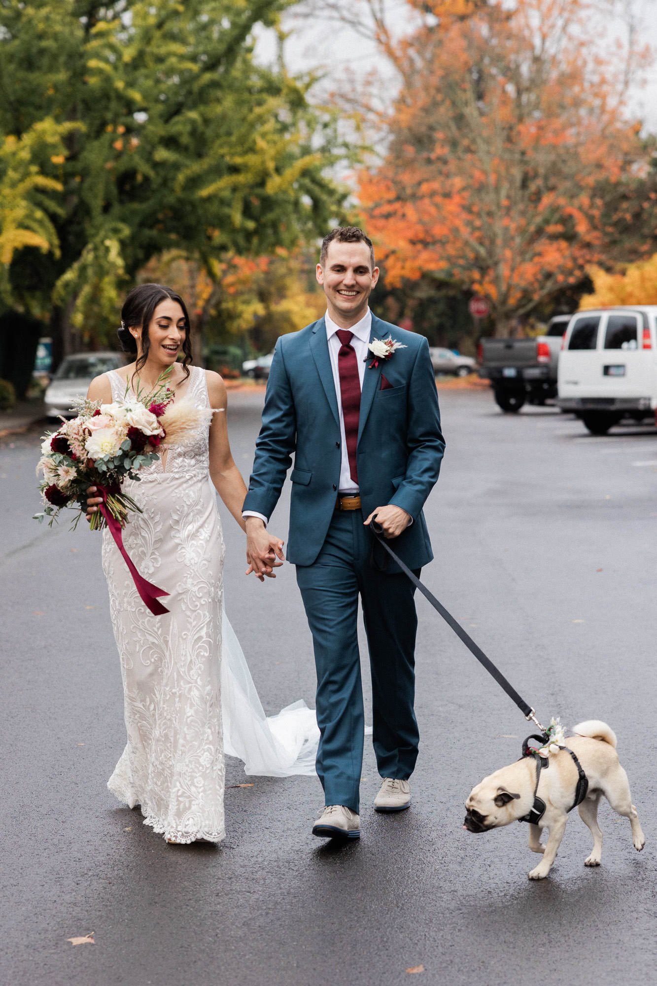 bride in white dress holding floral bouquet holds hand and walks with groom in blue suit and dog on leash
