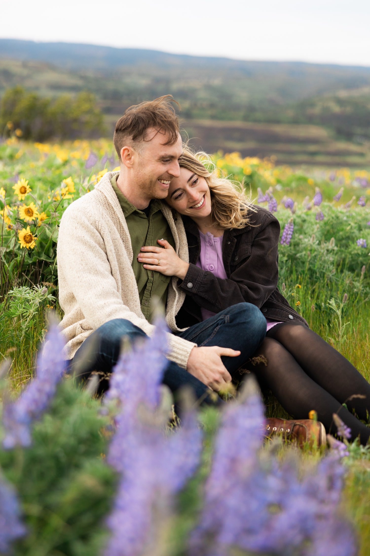 man and woman sit in field of balsamroot and lupine at rowena crest engagement session.jpg