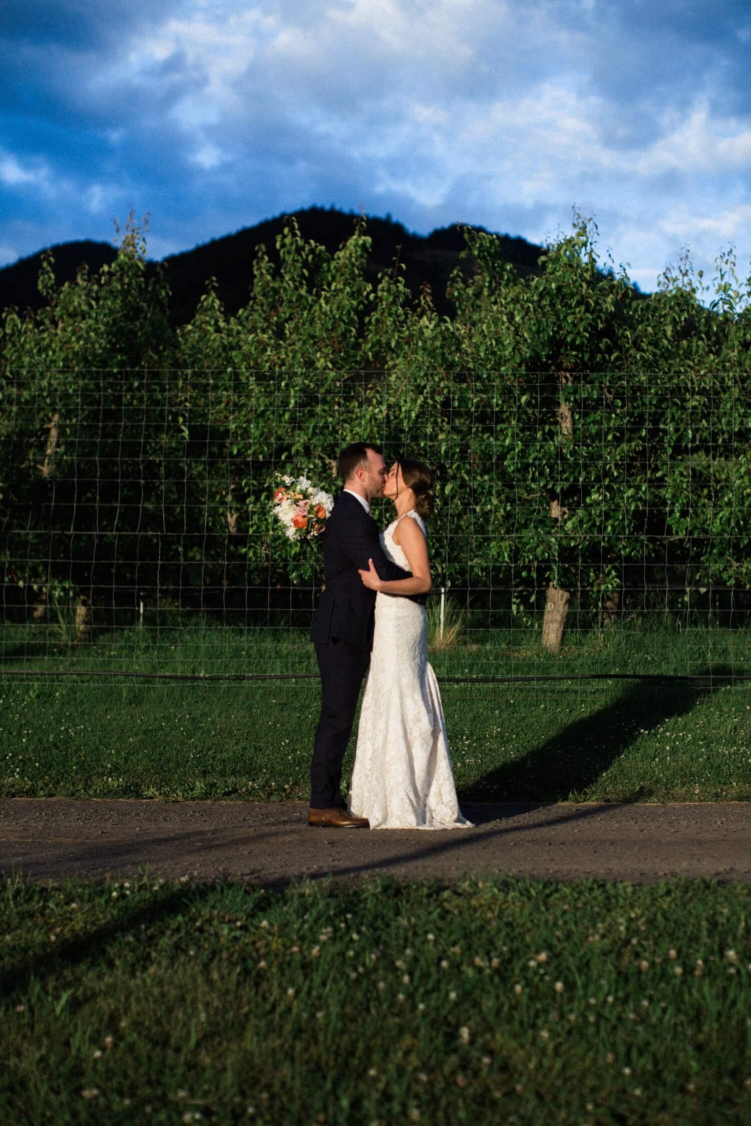 095_Mount Hood Organic Farms Wedding-Bride and groom hug and kiss each other in front of orchard trees.jpg