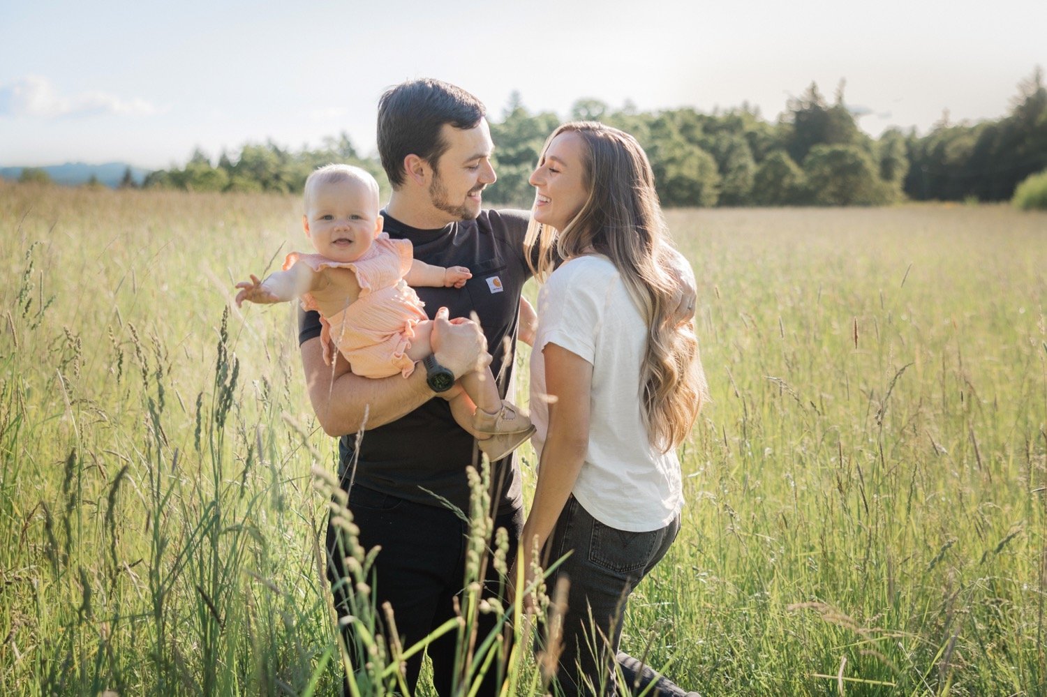 01_Portland Family Photo Session9_Woman in white shirt and man in black shirt hold baby in pink dress while standing in grassy field.jpg