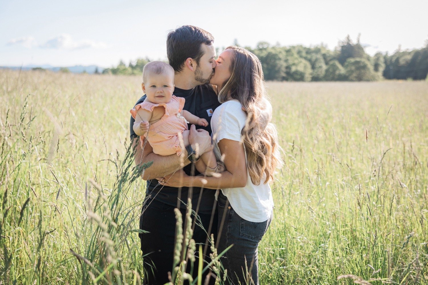 06_Portland Family Photo Session10_Woman in white shirt and kisses man in black shirt holding baby in pink dress in grassy field.jpg