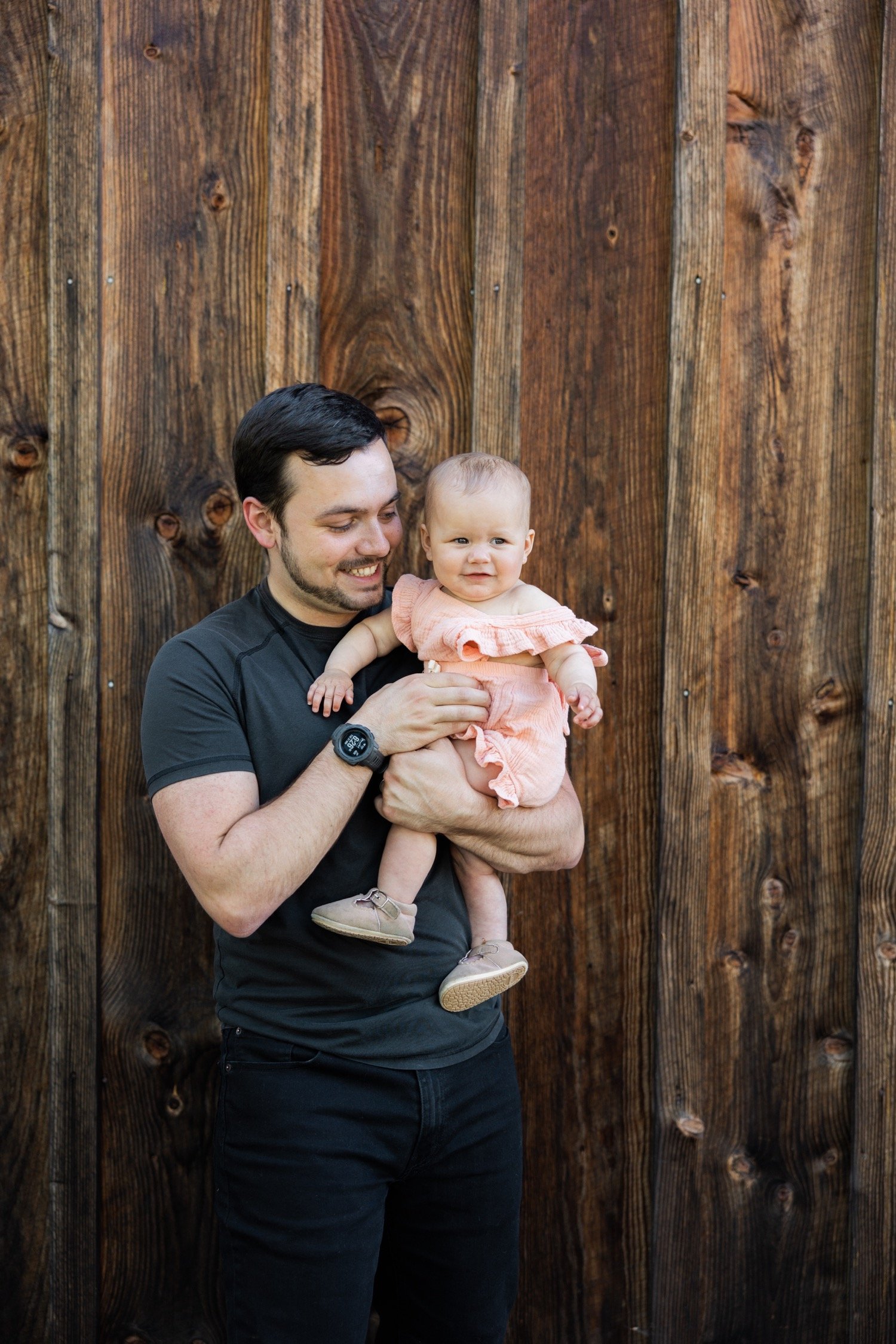17_Portland Family Photo Session20_Man in black shirt and black jeans holds baby in pink dress while standing against wood wall.jpg