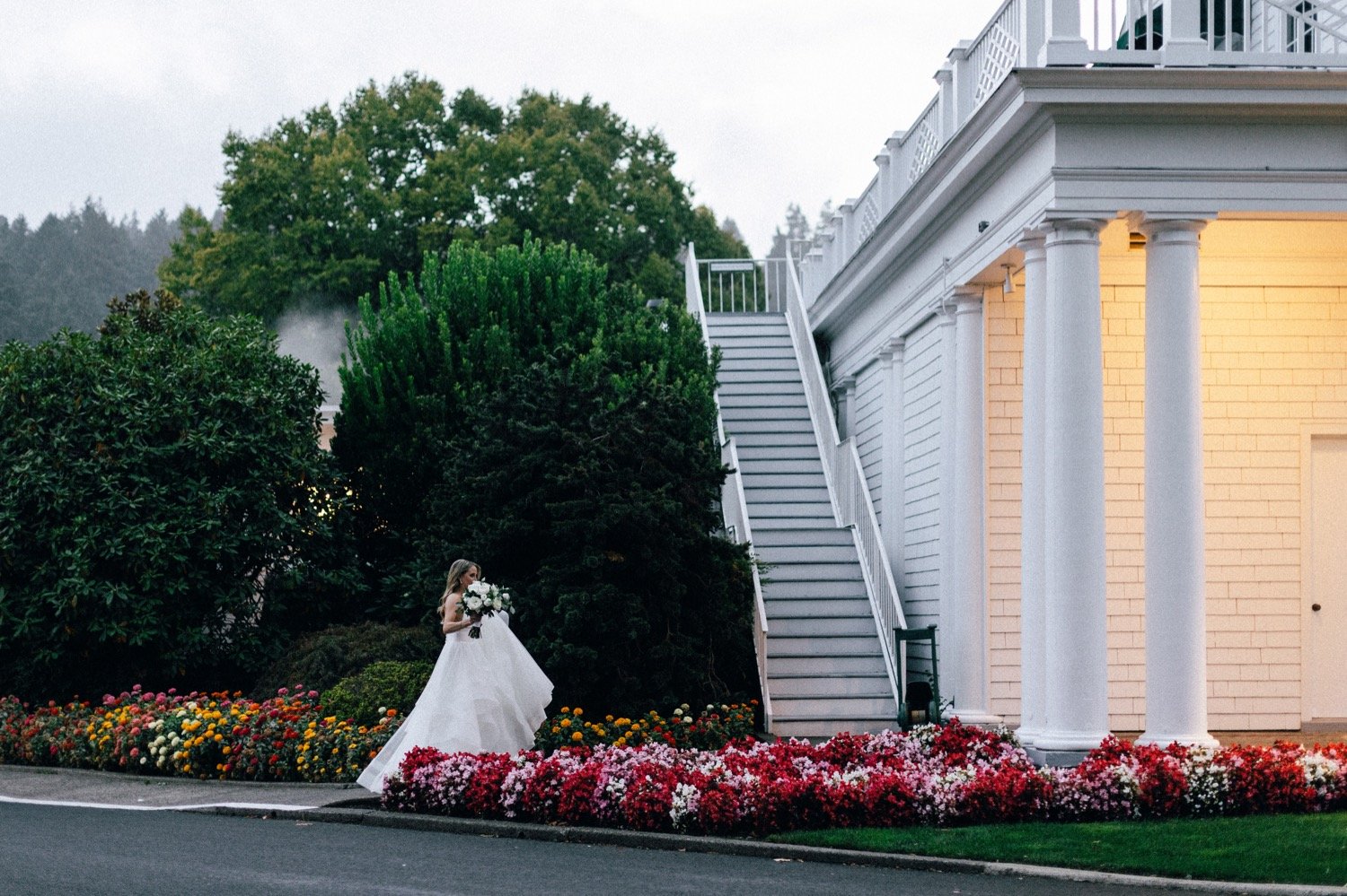  Bride in white dress holds flowers and dress while walking toward white building 