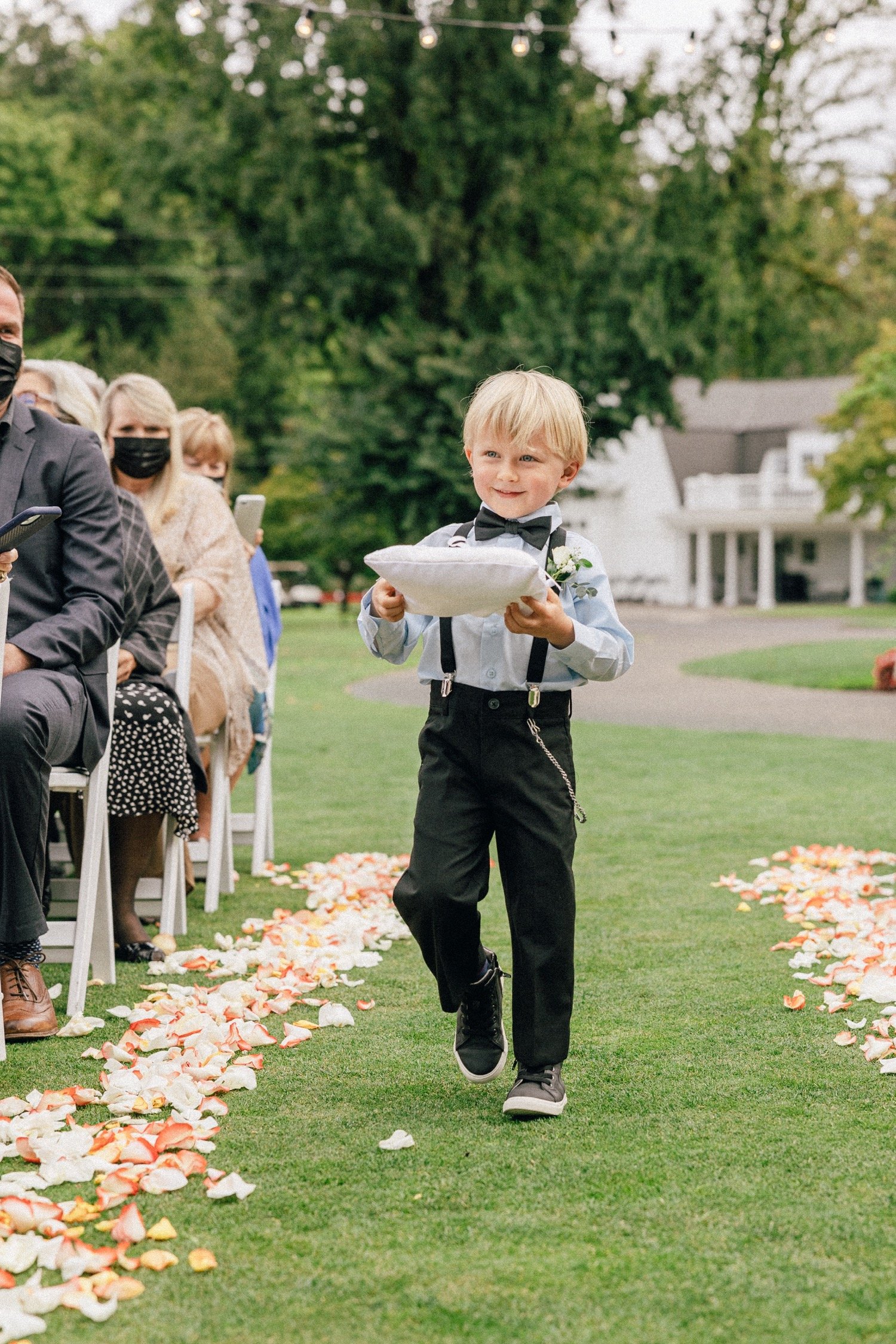  Ring bearer in blue shirt and black suspenders walks down aisle with pillow and rings 