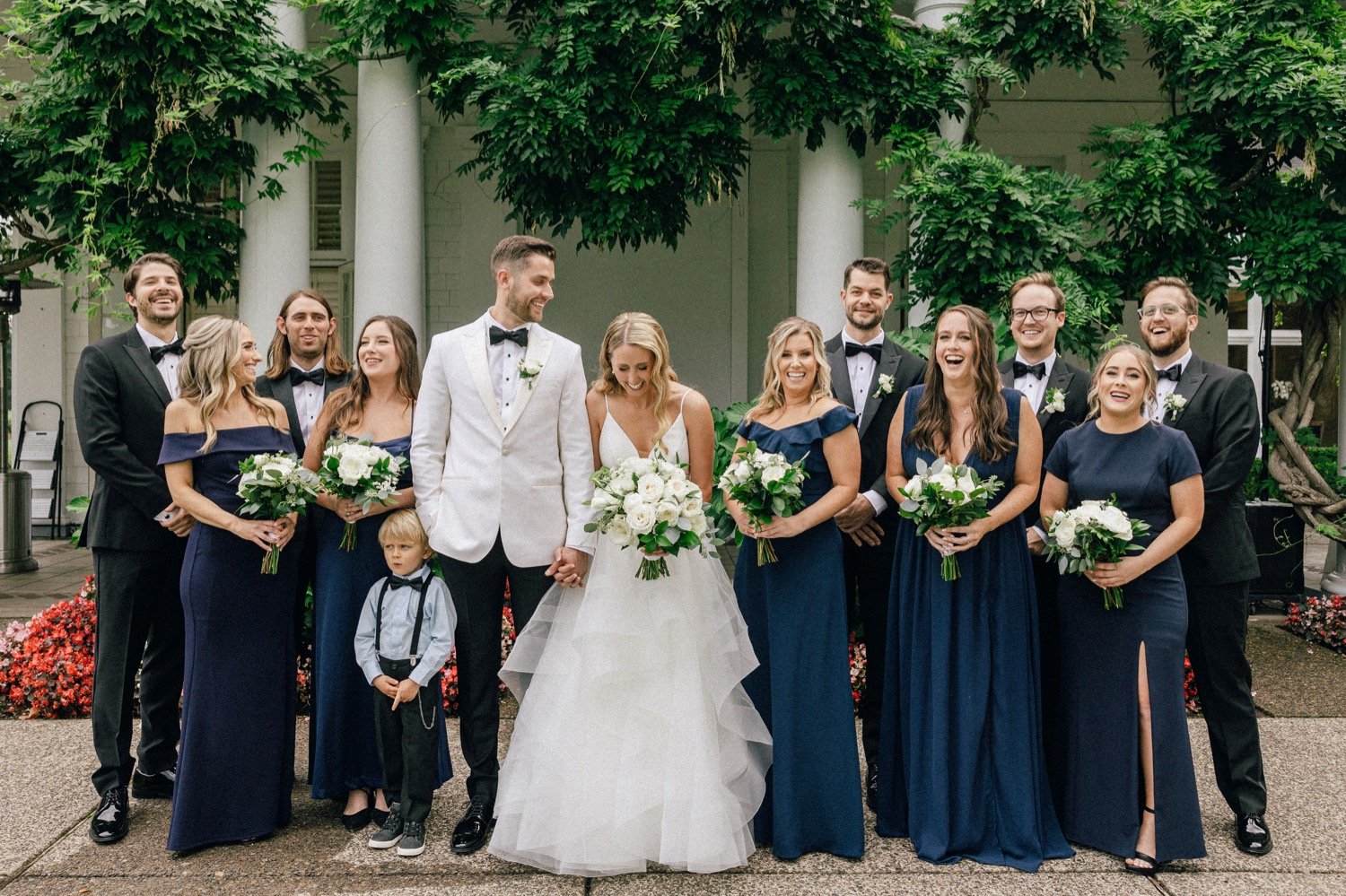  Wedding party laughs at one another while standing in front of white building and greenery at Waverley Country Club 