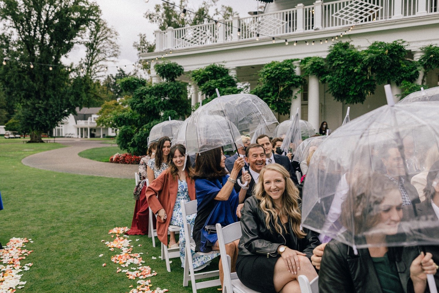  Wedding guests sit in white chairs and hold clear umbrellas during ceremony 