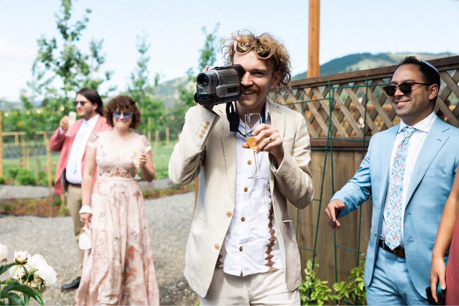 021_The Orchard Hood River Wedding57_man in khaki suit and white shirt holds camcorder and glass of wine.jpg