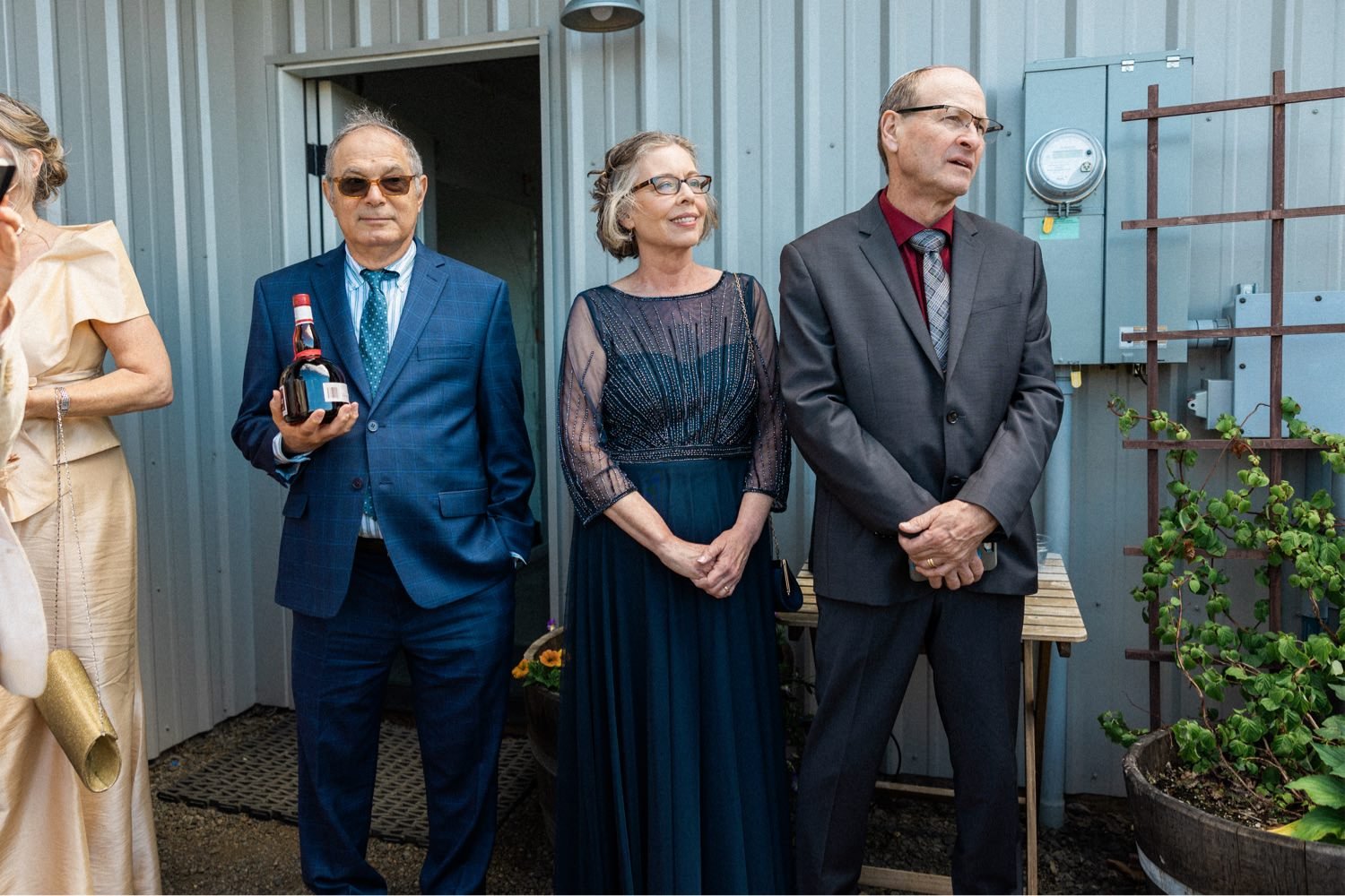 024_The Orchard Hood River Wedding58_man in gray suit and woman in navy dress stand next to man in navy dress holding bottle of liquor.jpg