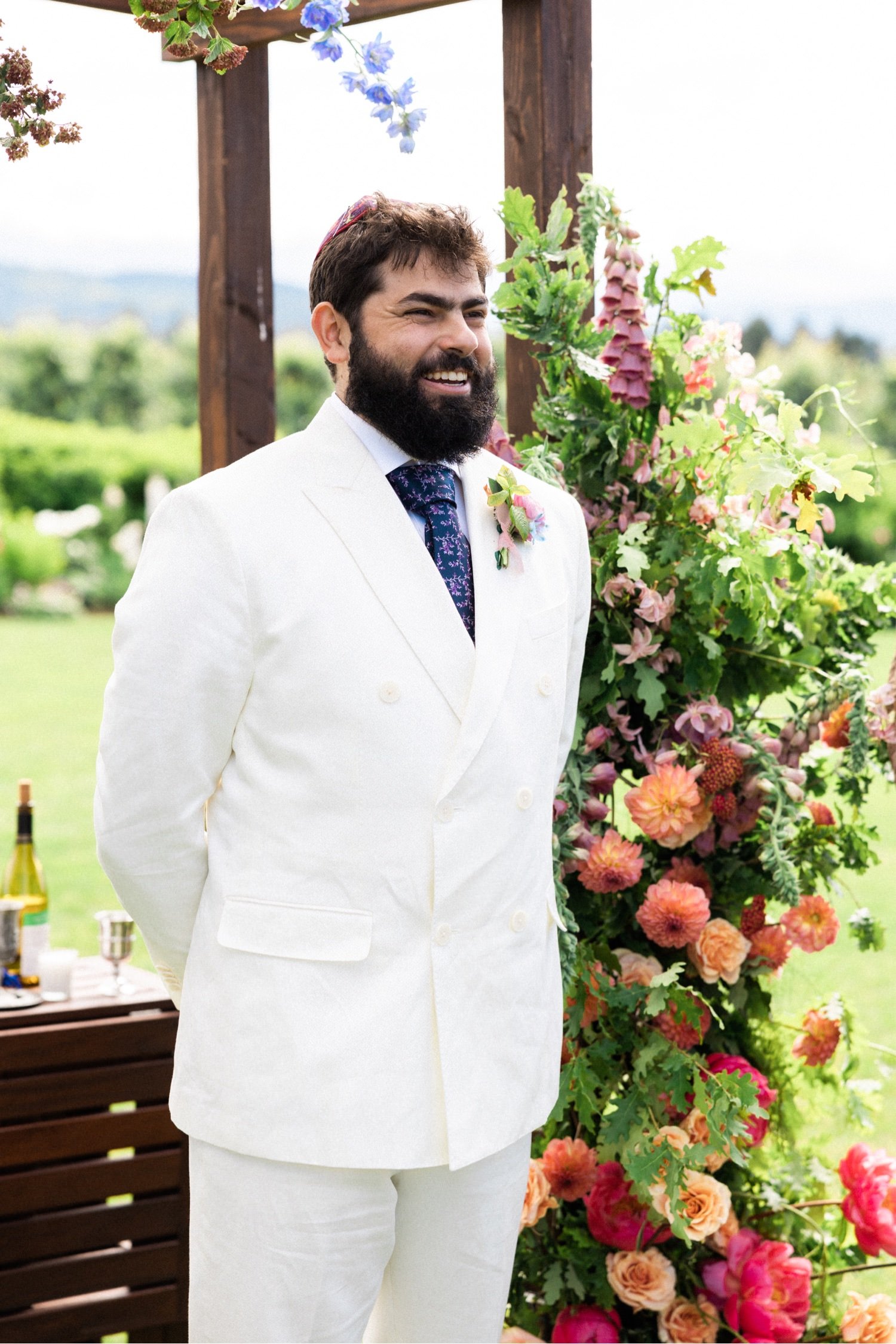 038_The Orchard Hood River Wedding83_groom wearing white suit and kippah sands under floral arbor and smiles.jpg