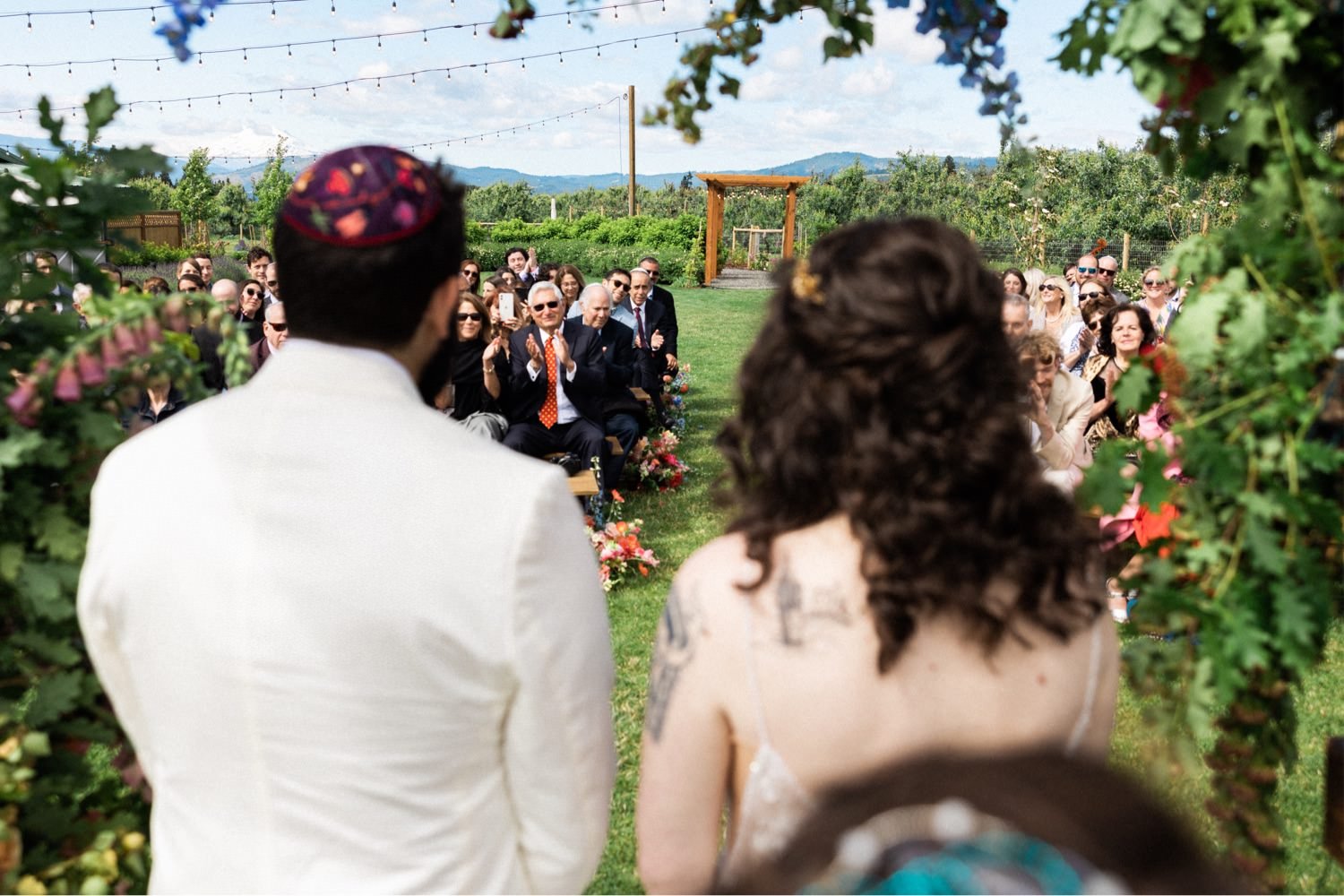 043_The Orchard Hood River Wedding89_bride and groom wearing kippah look out at wedding guests during ceremony.jpg