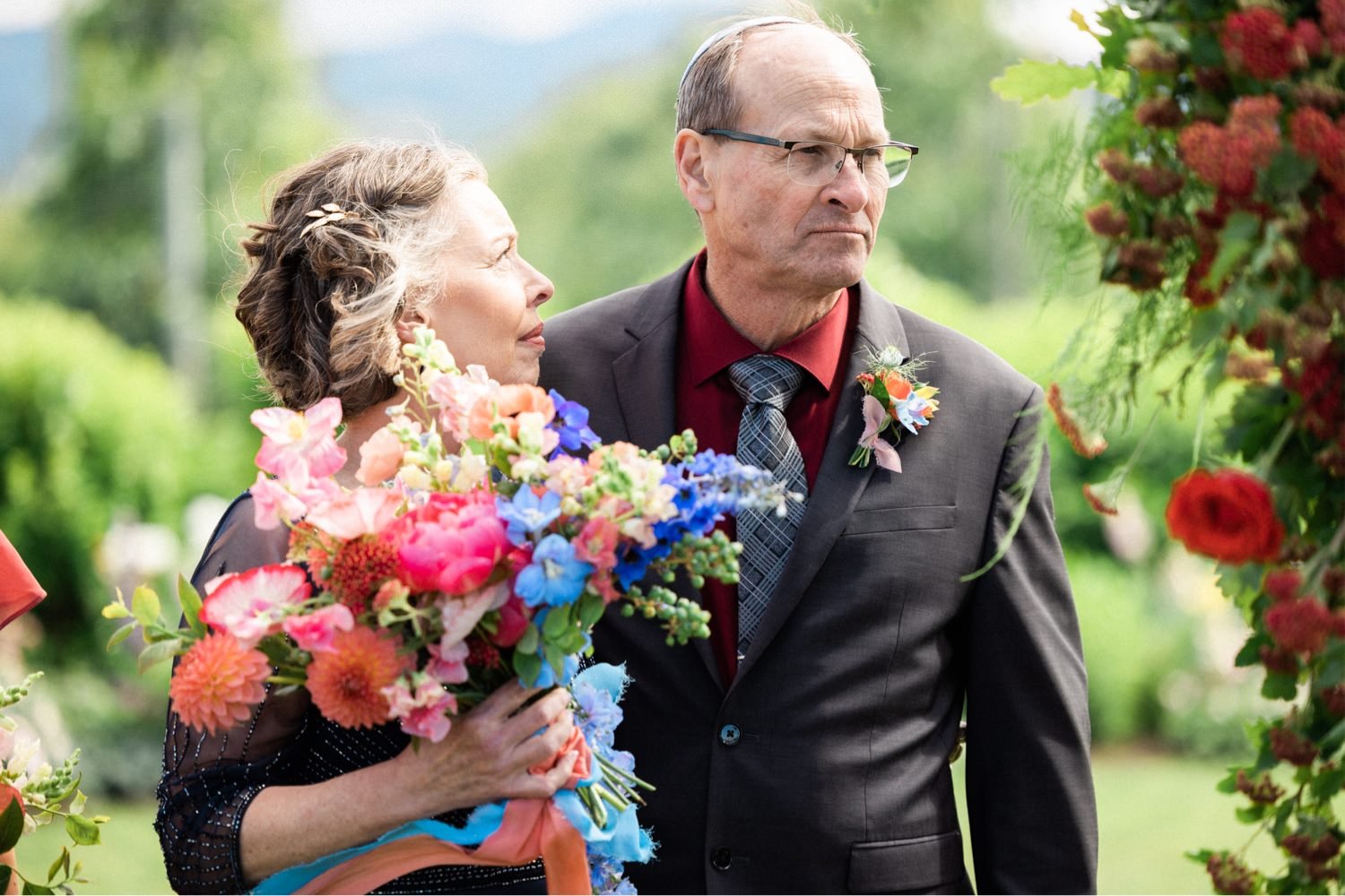 049_The Orchard Hood River Wedding5_woman holding bouquet of pink and orange flowers looks toward man in black suit.jpg