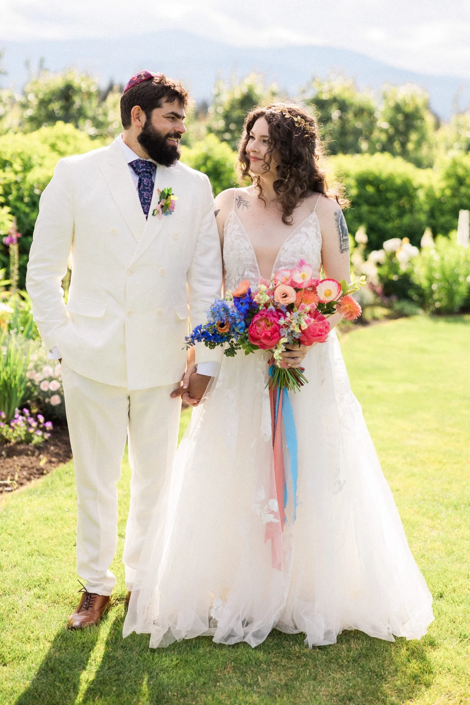 072_The Orchard Hood River Wedding115_bride in white dress holding bouquet of purple and pink flowers holds hand of groom in white suit and kippah.jpg