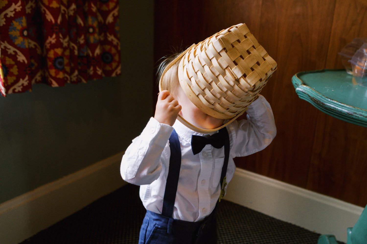  toddler in suspenders and bowtie puts wicker basket over his head 