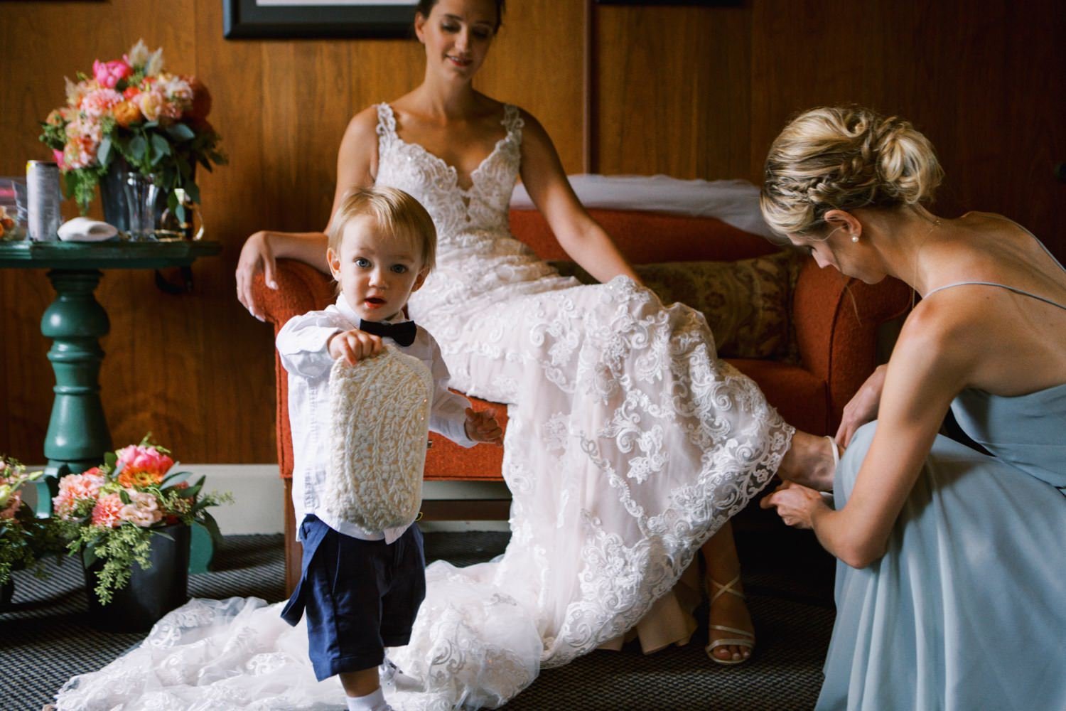  bride sits on couch as bridesmaid helps put on her shoes while little boy stands in front of them 