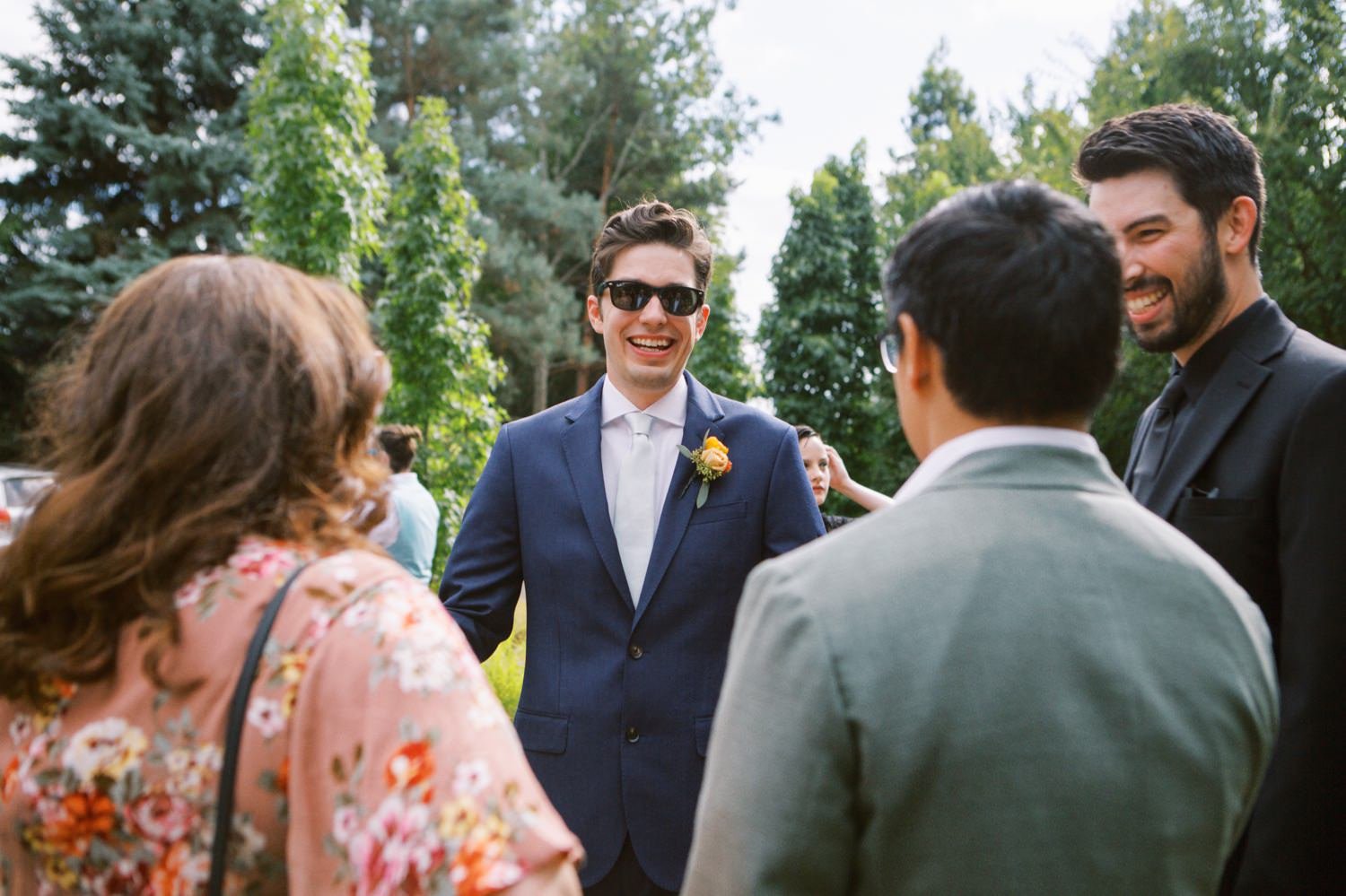  groom in navy blue tux and sunglasses is greeted by three people 