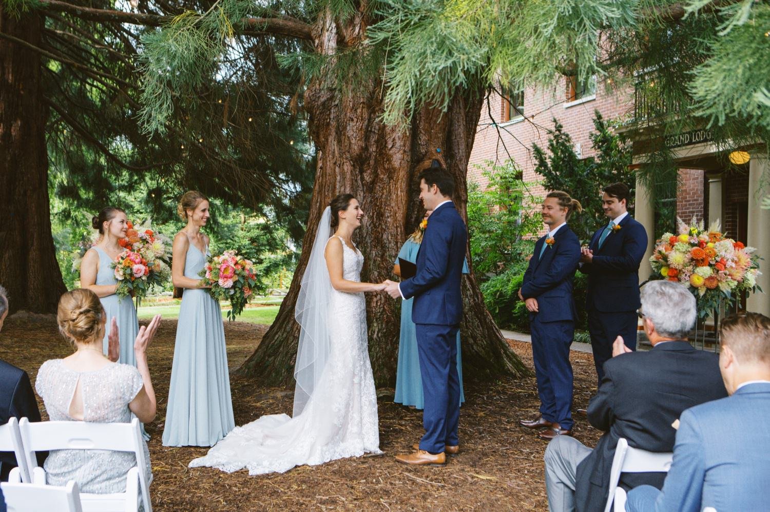  bride in white dress and bridesmaids in light blue dresses stand next to groom and two groomsmen in navy suits during wedding ceremony at mcmenamins grand lodge 
