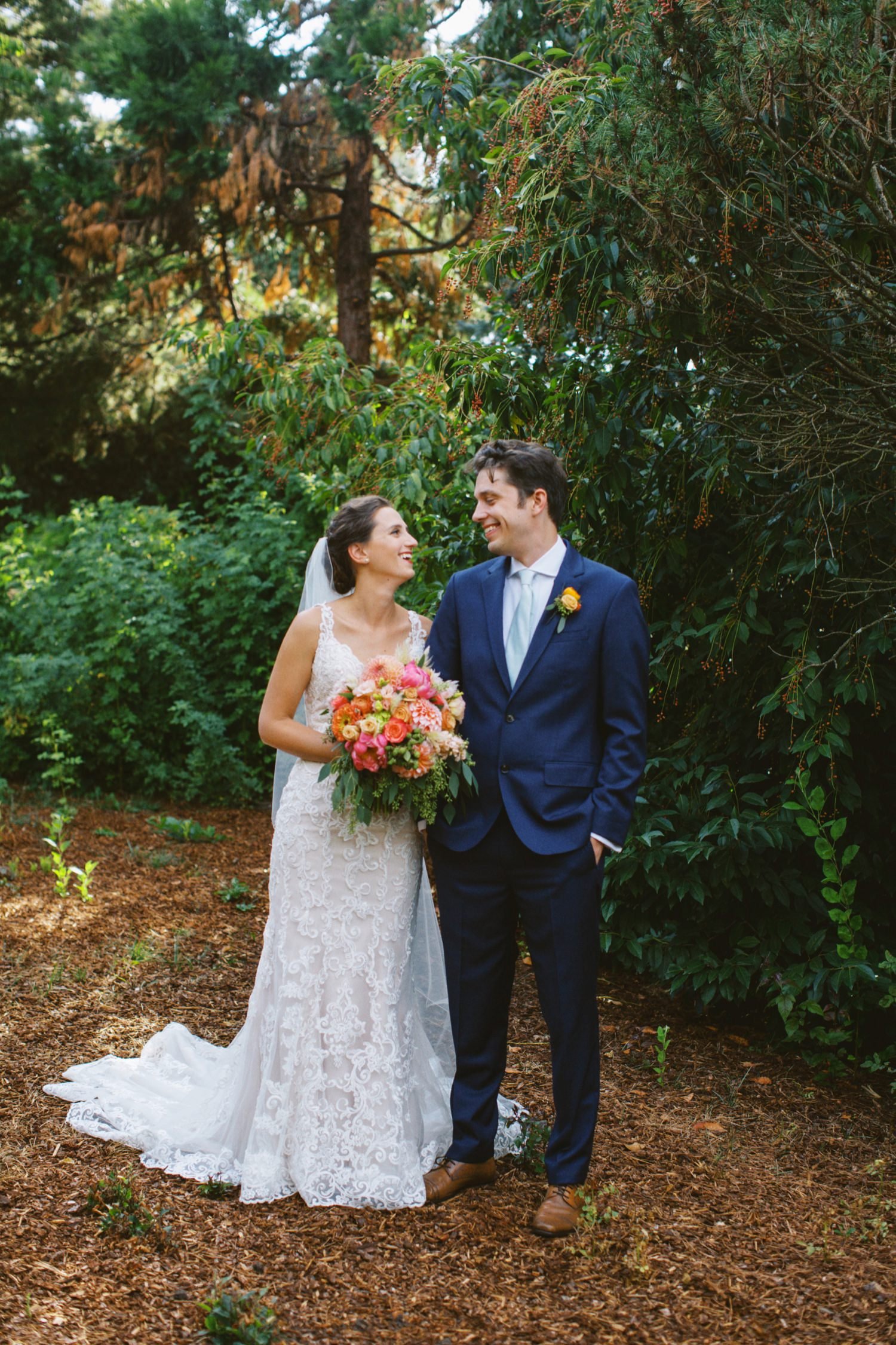  bride in white dress holding bouquet of pink and orange flowers stands next to and smiles at groom in navy blue suit 