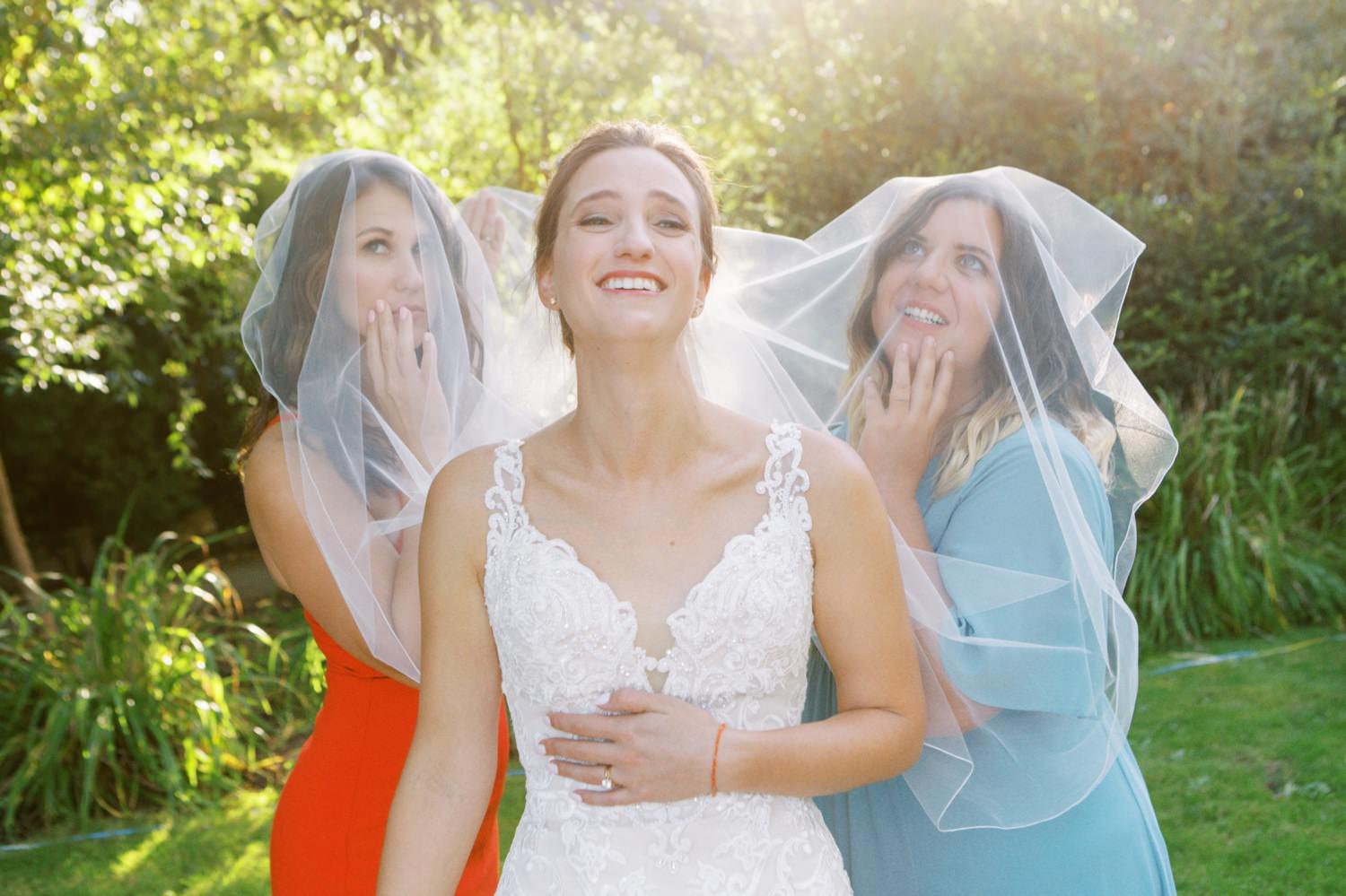  two women stand behind smiling bride and put veil over their faces 